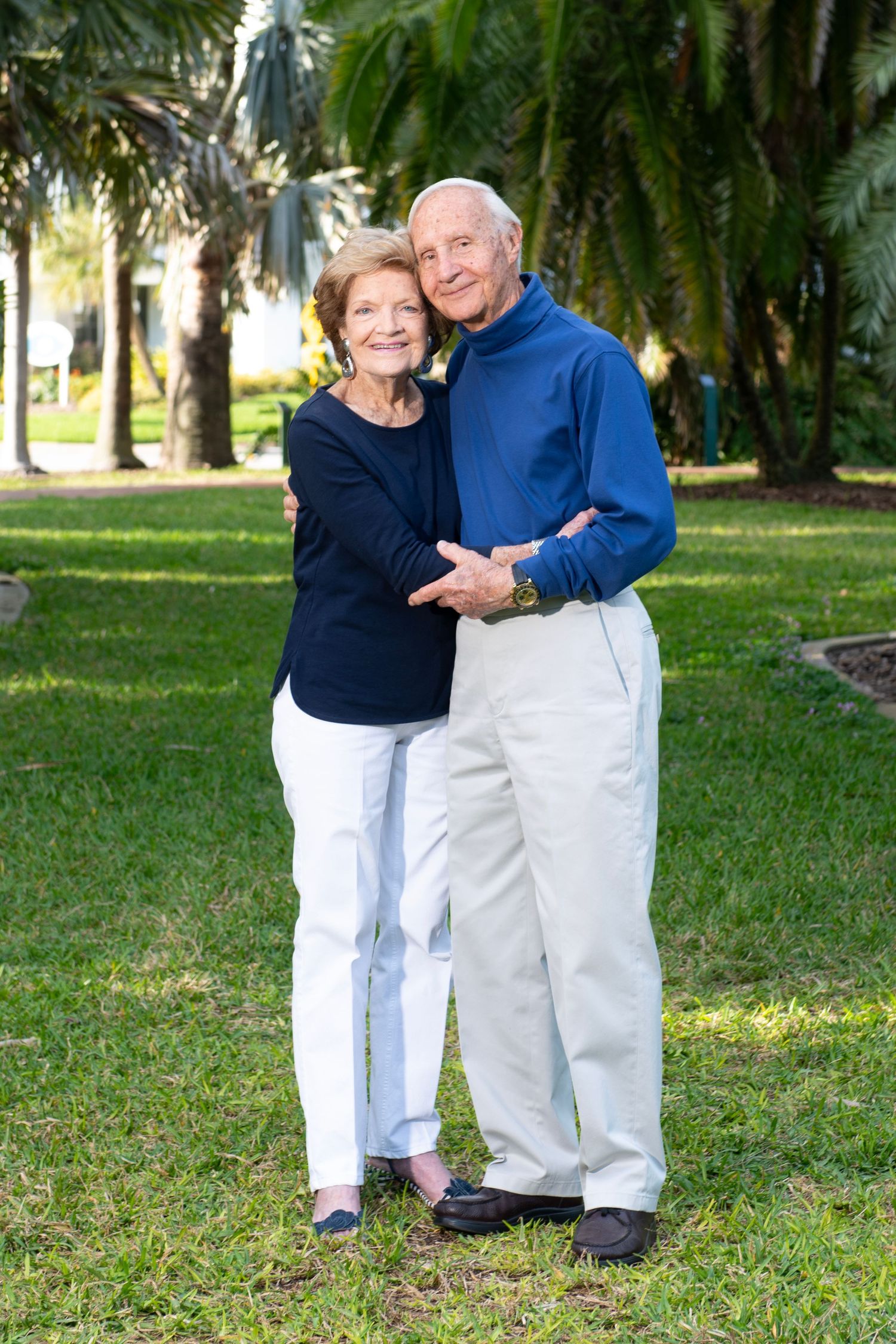 Happy senior couple embracing outdoors on green lawn with tropical palm trees in background.