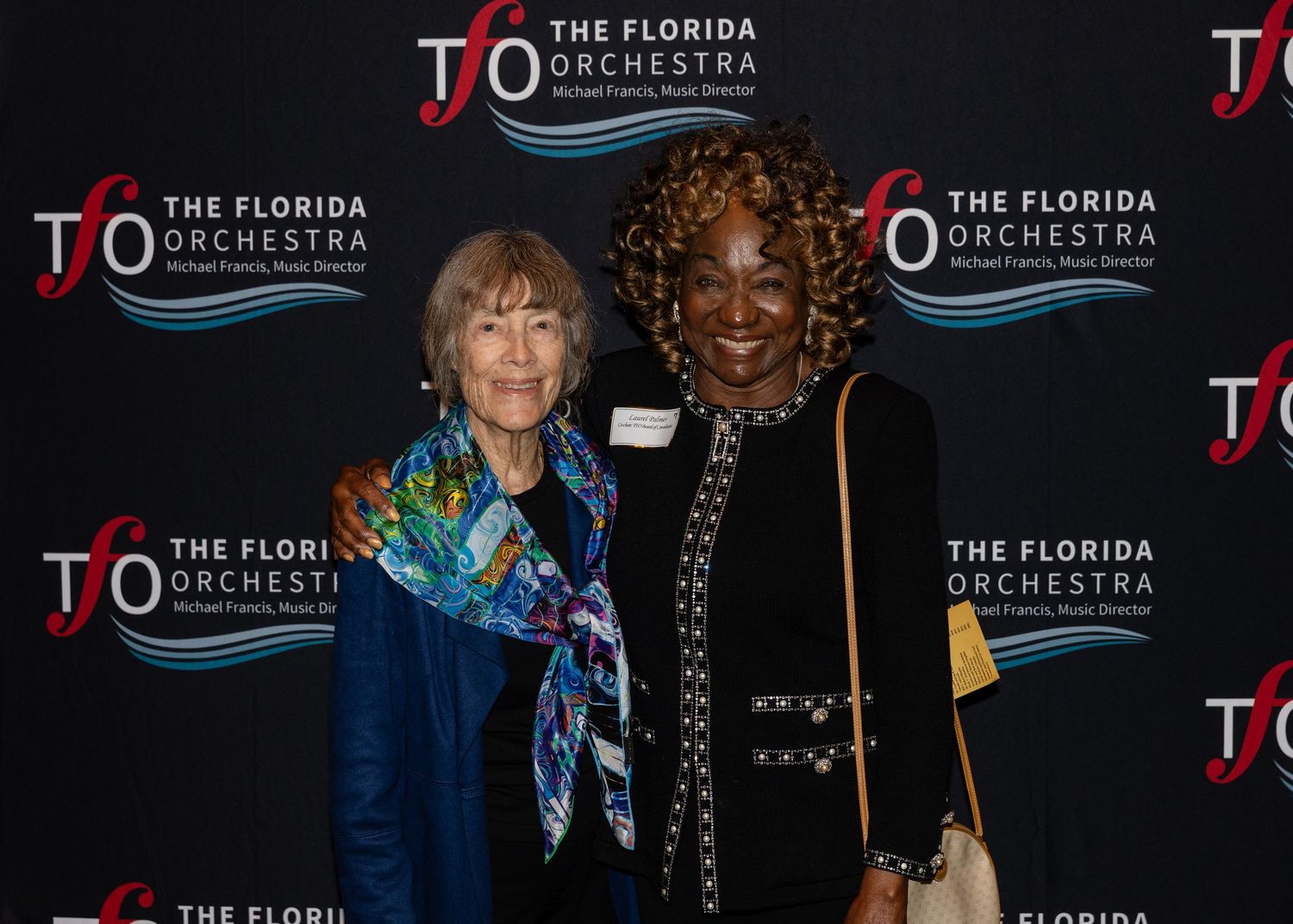 Two women pose together at a Florida Orchestra event, smiling in front of branded backdrop signage.