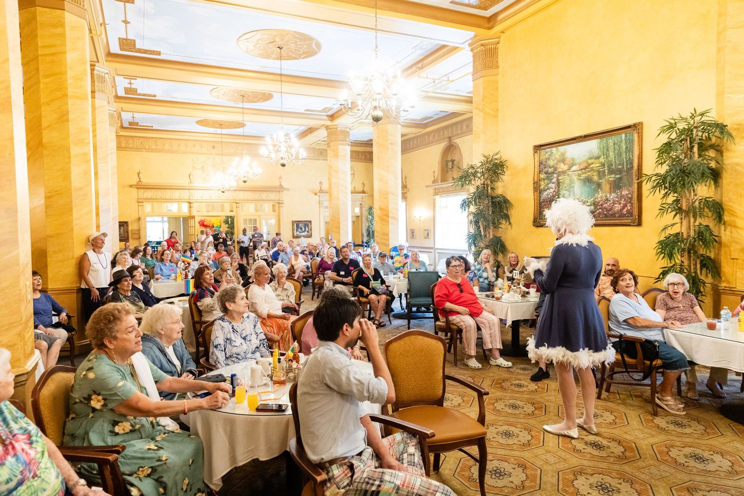 Speaker addresses a large crowd of seniors at a Pride celebration in a grand ballroom.
