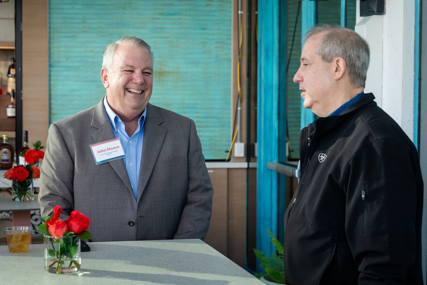 Two men in business casual attire share a laugh while conversing at a counter with red roses nearby.