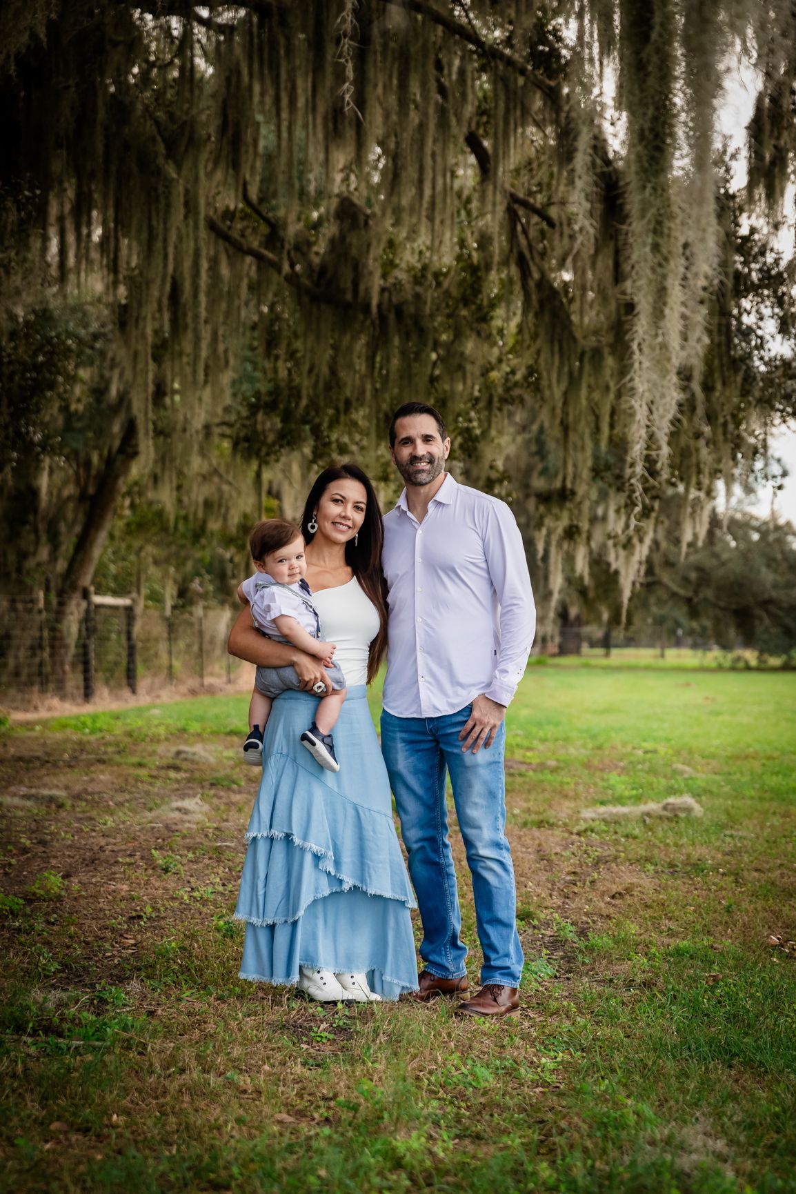 Family portrait of a couple with baby under Spanish moss-draped oak trees on a green lawn.