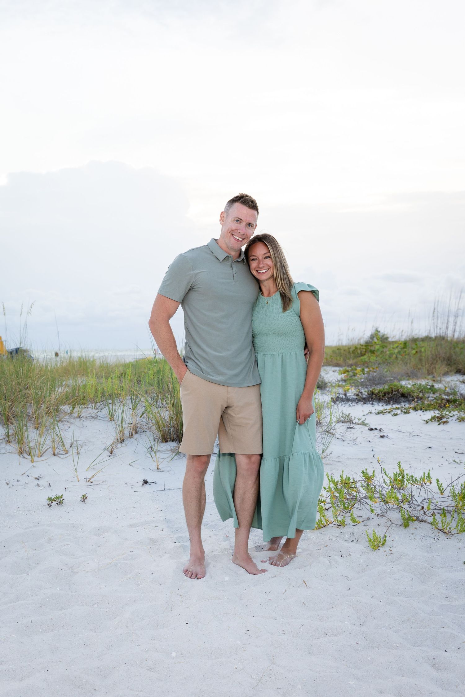 Happy couple posing together on sandy beach, woman in teal maxi dress, man in green polo.