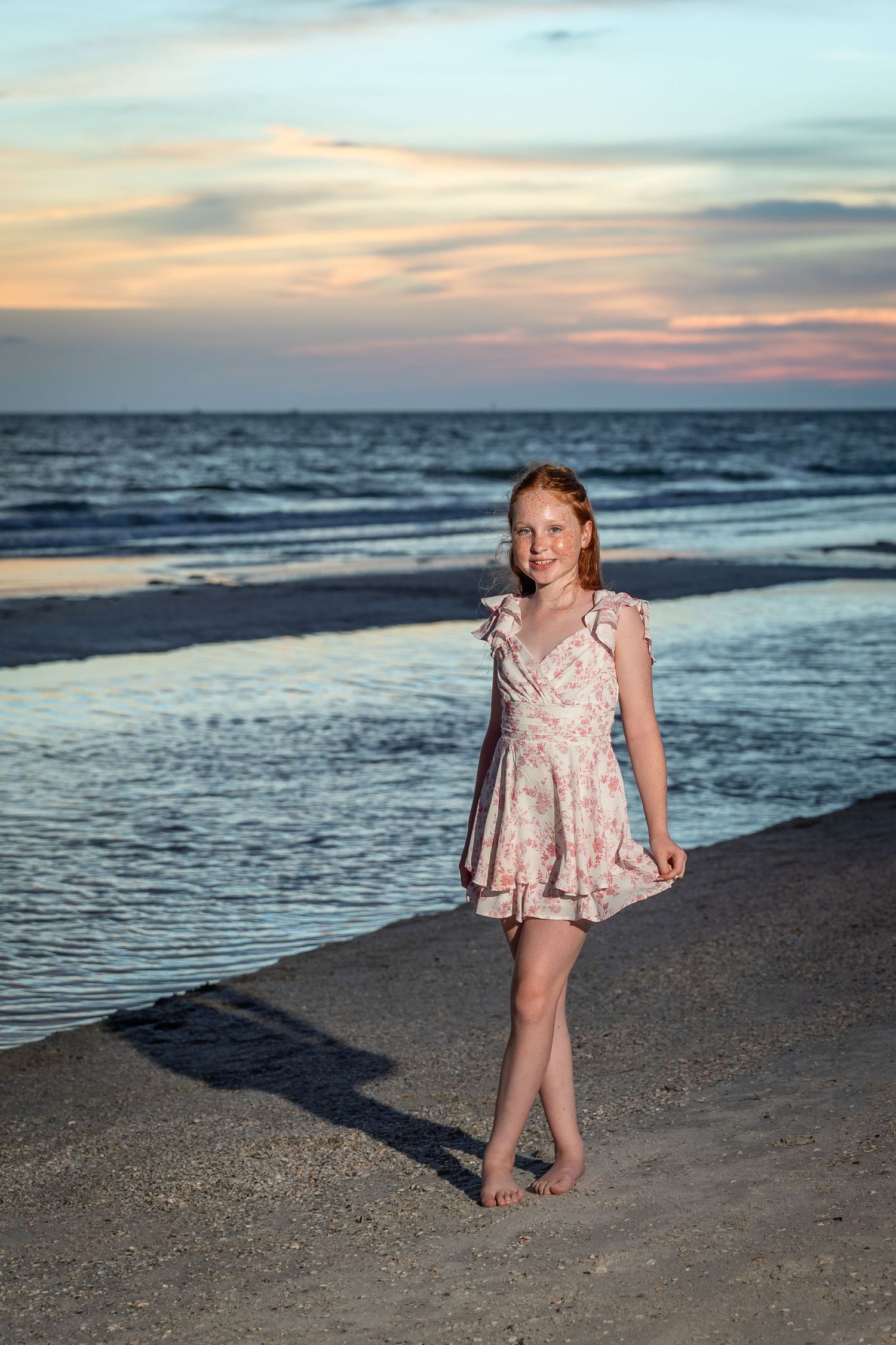 Young redhead girl in floral dress standing barefoot on sandy beach at sunset with ocean waves behind her.