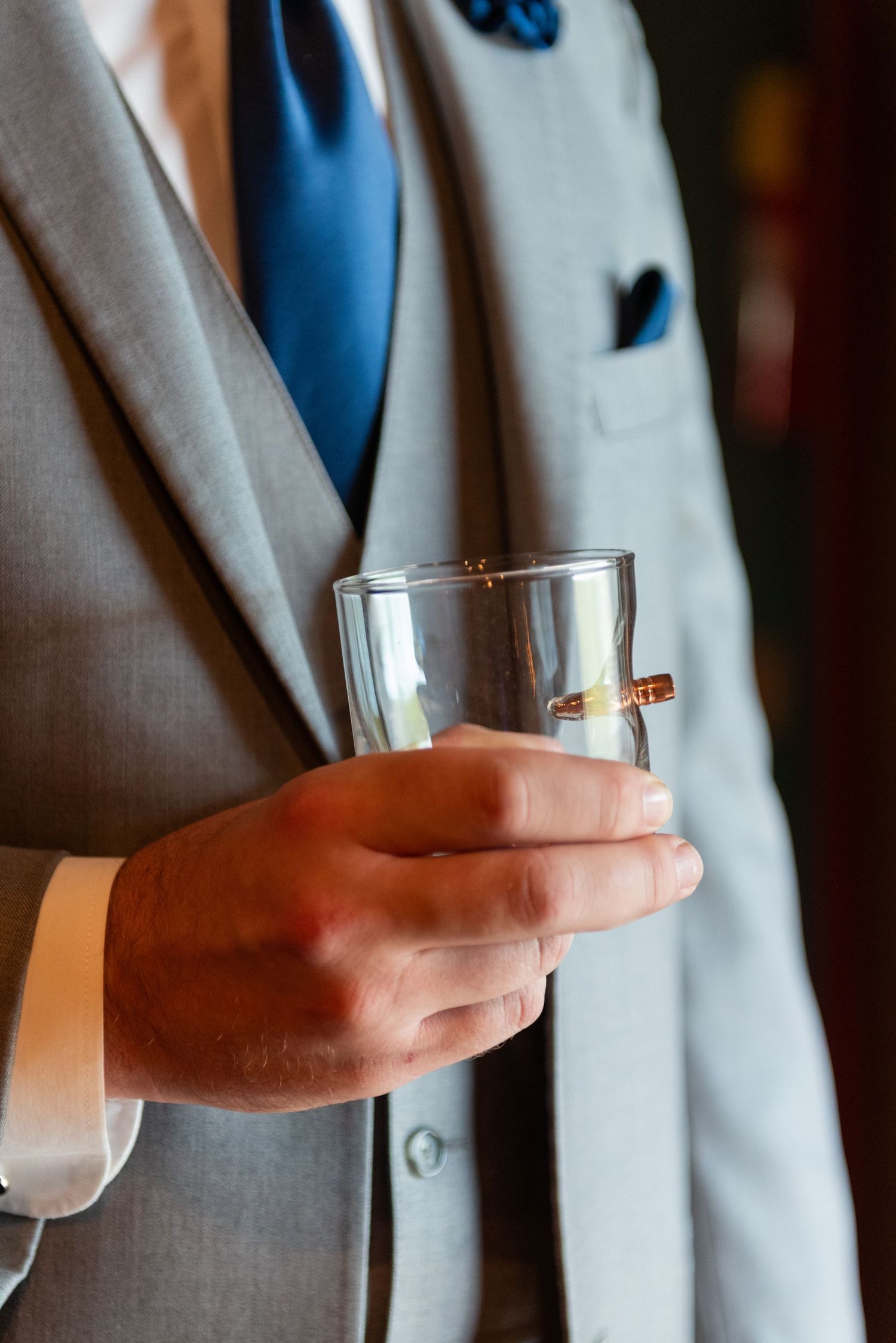 Groom in gray suit holds whiskey glass at wedding reception.