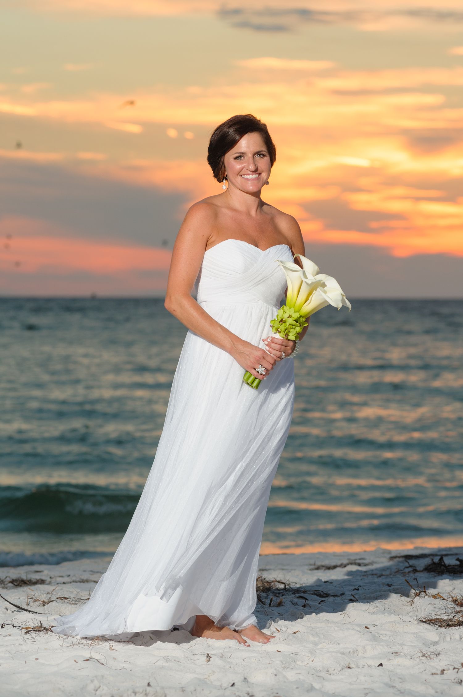 Radiant bride in white strapless gown holding calla lily bouquet at sunset beach wedding.
