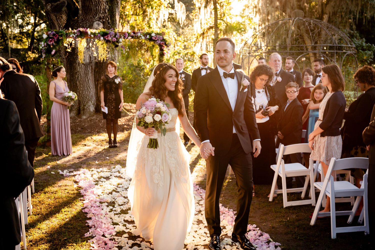Happy newlyweds walk down petal-lined aisle after outdoor wedding ceremony with guests cheering.