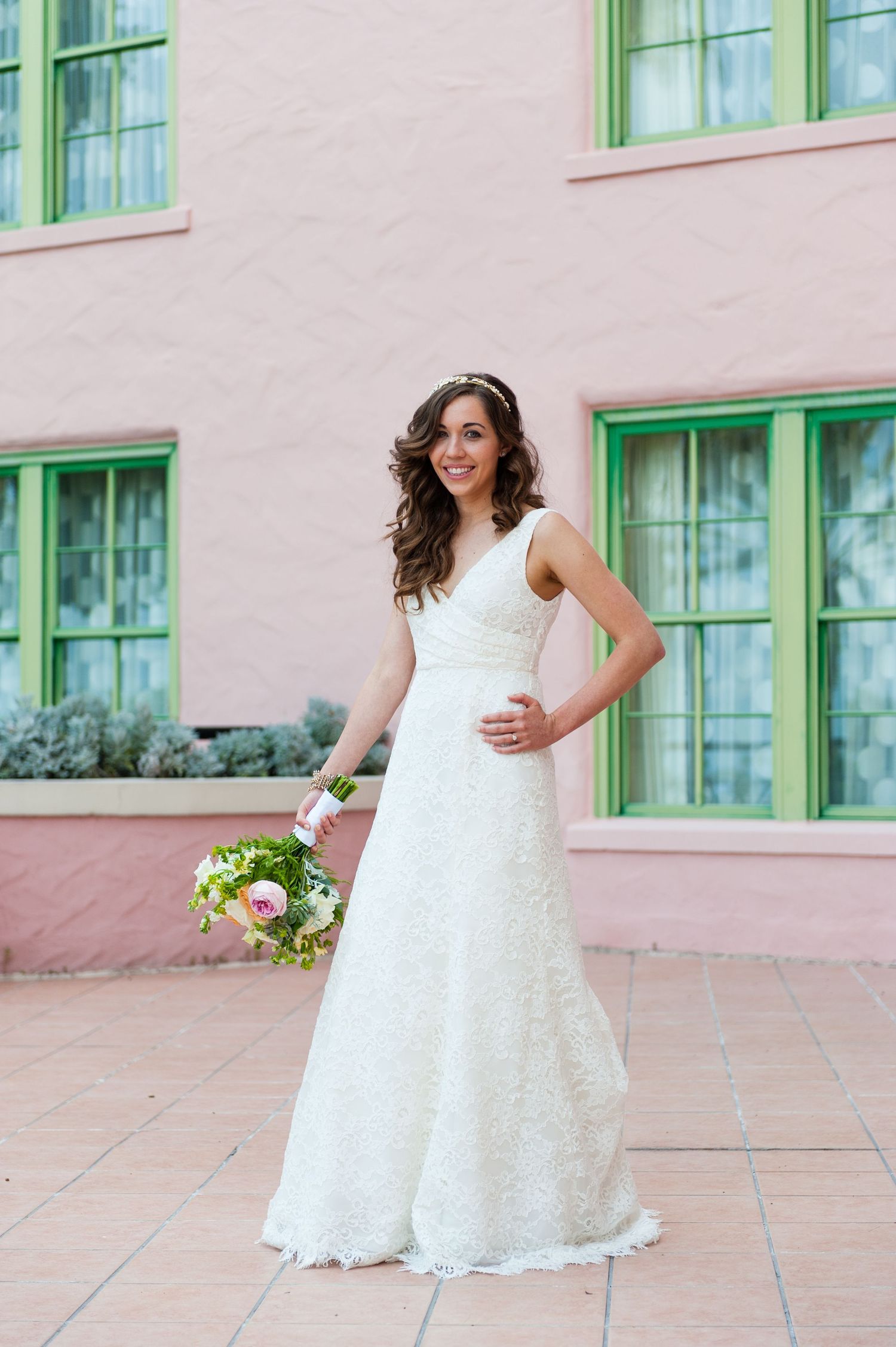 Beautiful bride in a lace wedding gown holding a bouquet outside a pink building.
