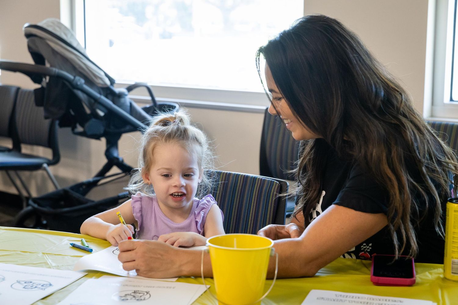 A woman helps a young girl with an art activity at a yellow table in a bright classroom setting.