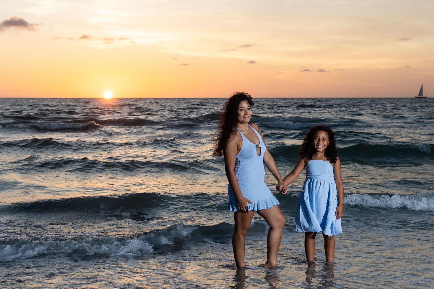Mother and daughter holding hands in blue dresses stand in ocean waves during a stunning golden sunset.
