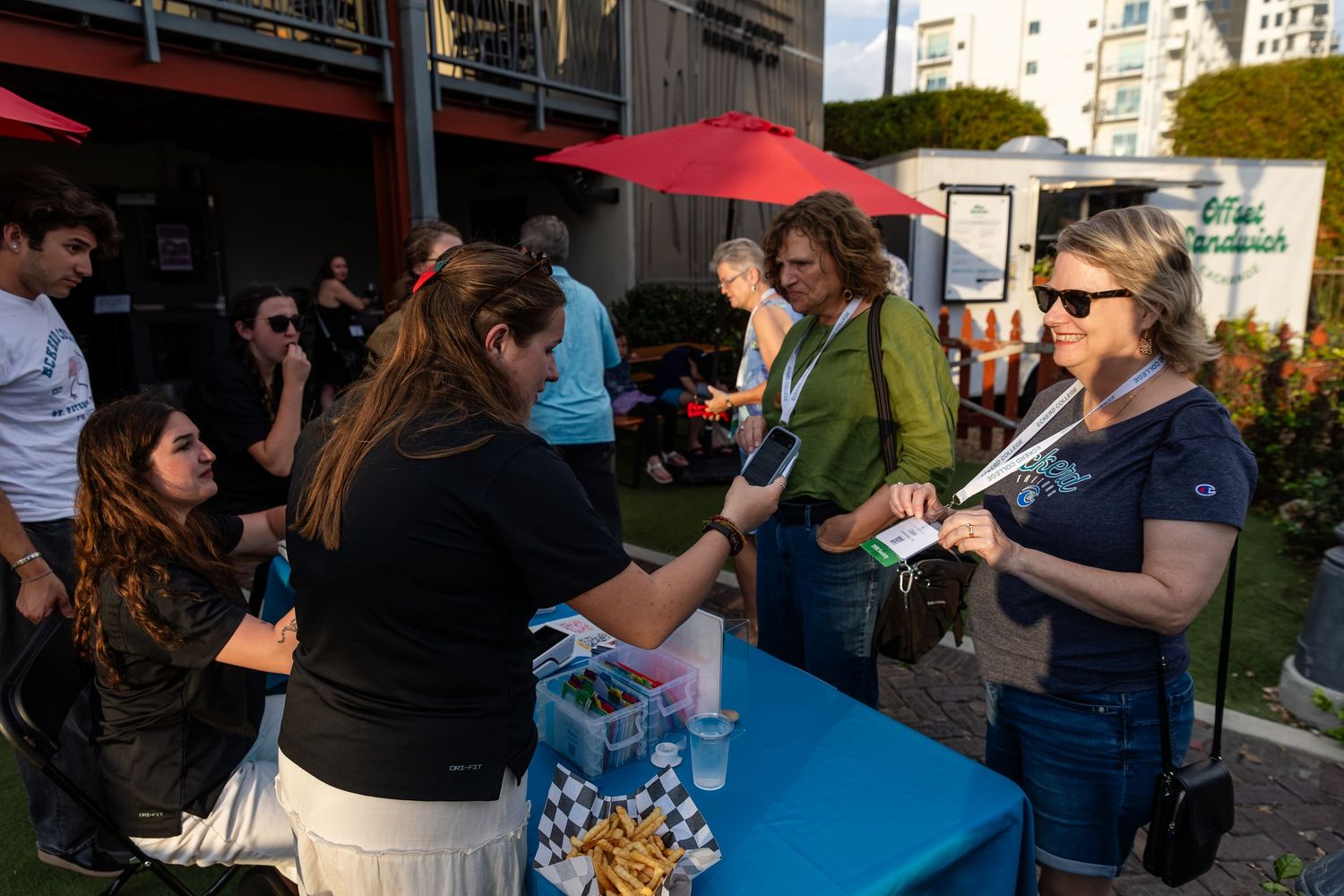 People networking at an outdoor event with a blue table, red umbrellas, and food truck in the background.
