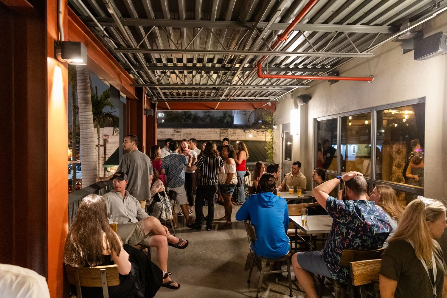 Busy indoor-outdoor bar patio with groups of people socializing at wooden tables under exposed ceiling beams at night.