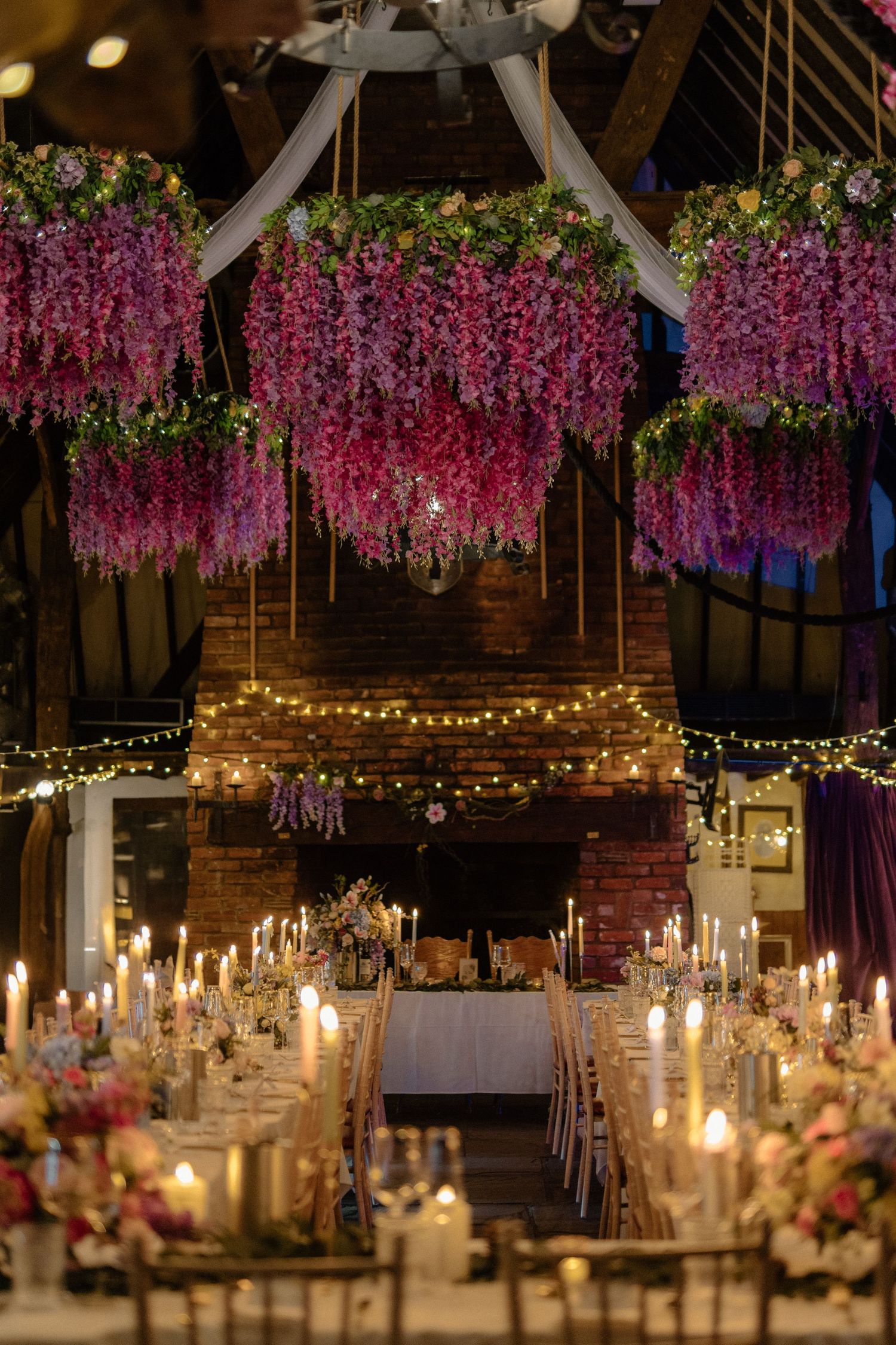 An elegant wedding reception hall with hanging floral chandeliers, candlelit tables, and fairy lights.