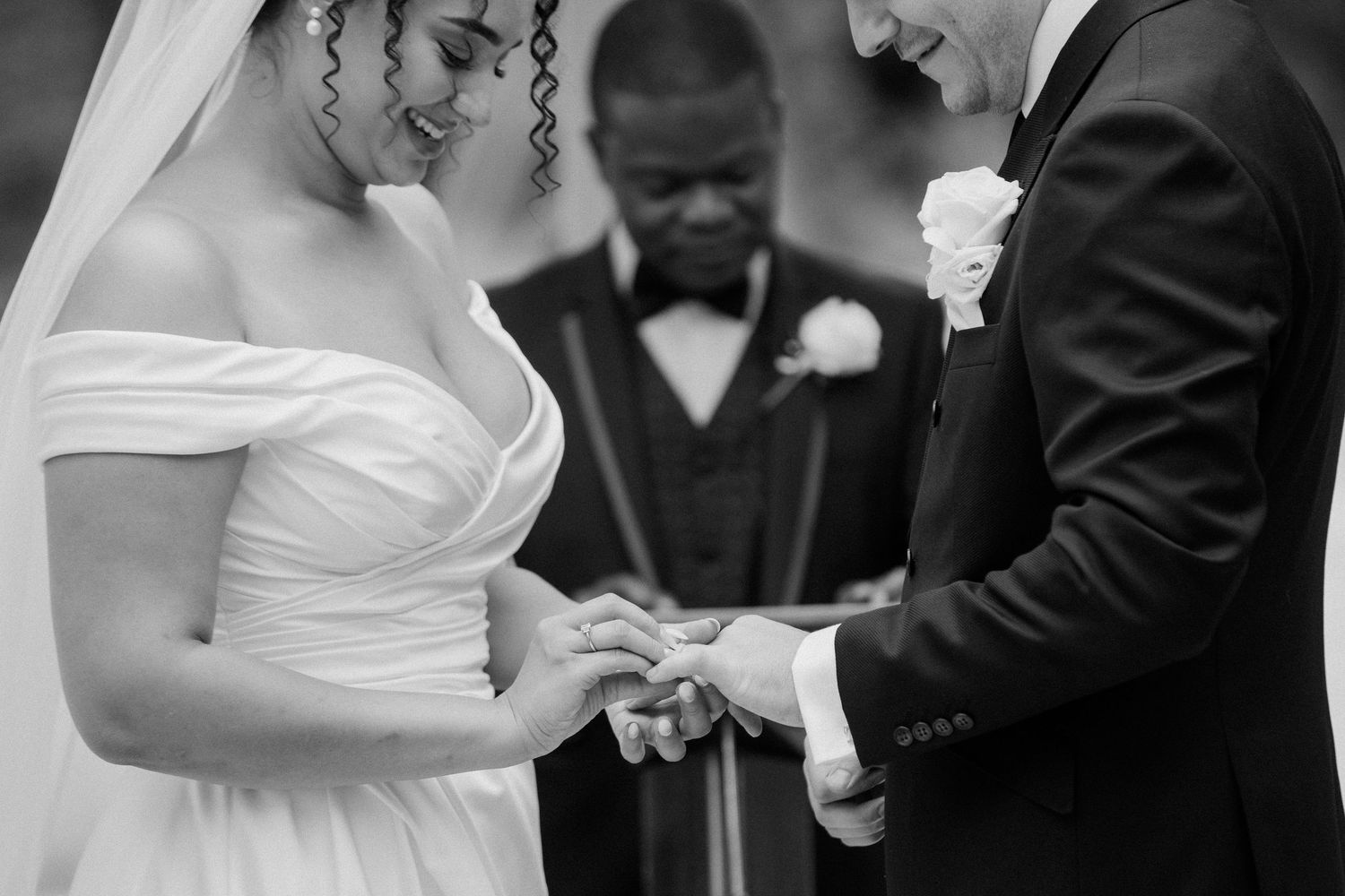 Black and white photo of couple exchanging wedding rings with officiant during outdoor ceremony.