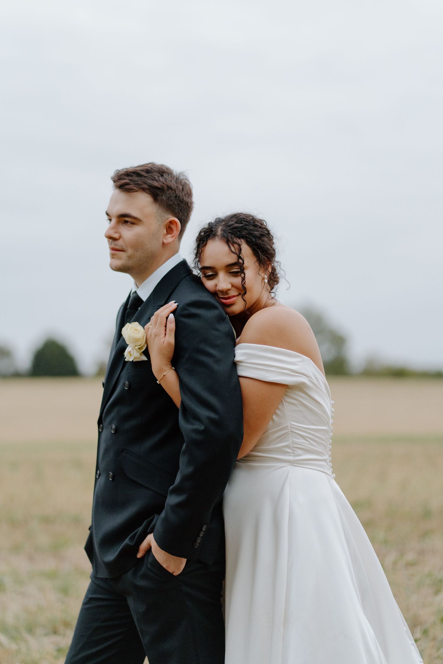 Bride leans lovingly against groom's back as they pose together in an open countryside field.