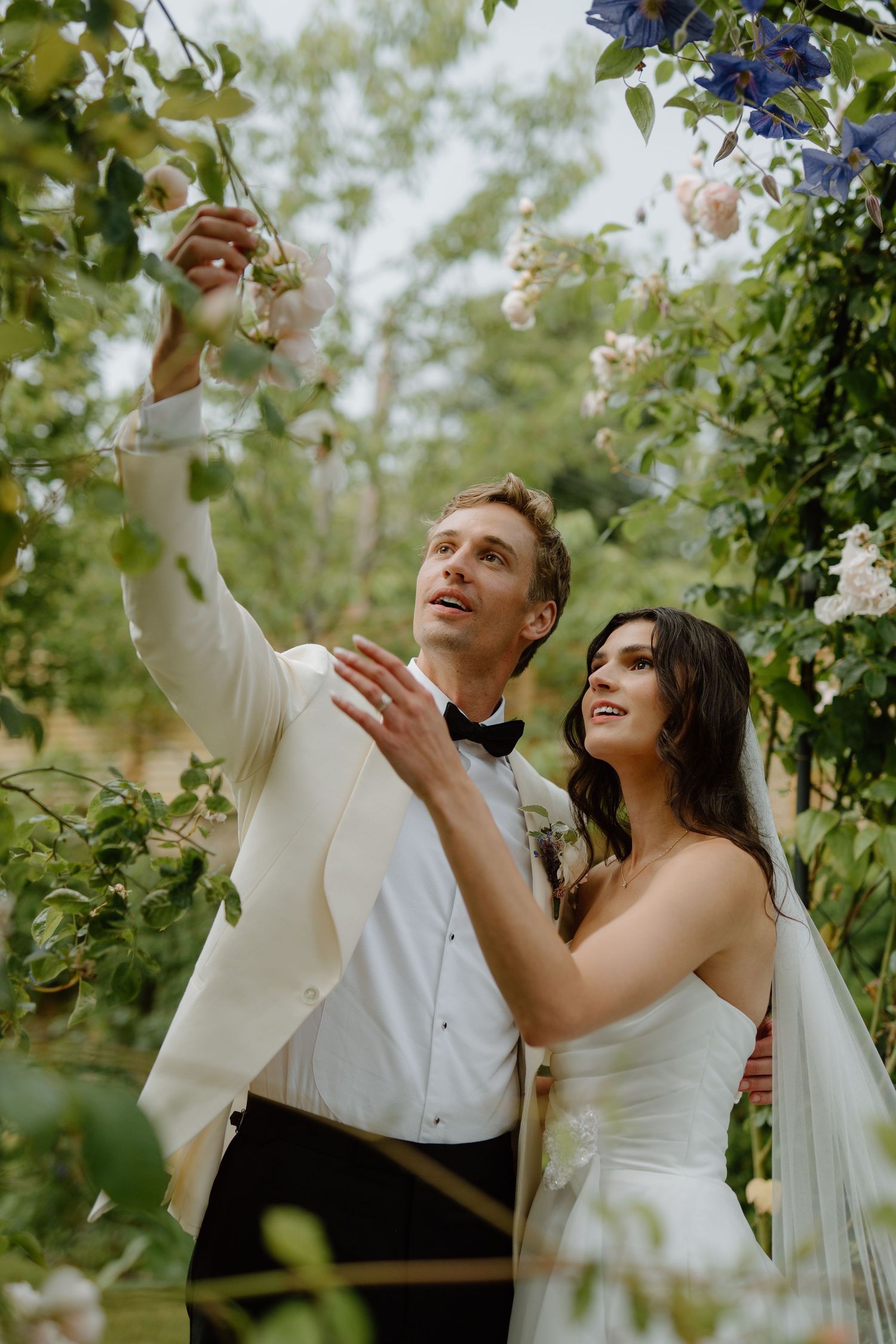 Bride and groom admire blooming roses on a floral arch, groom in white tuxedo reaching up to touch flowers.