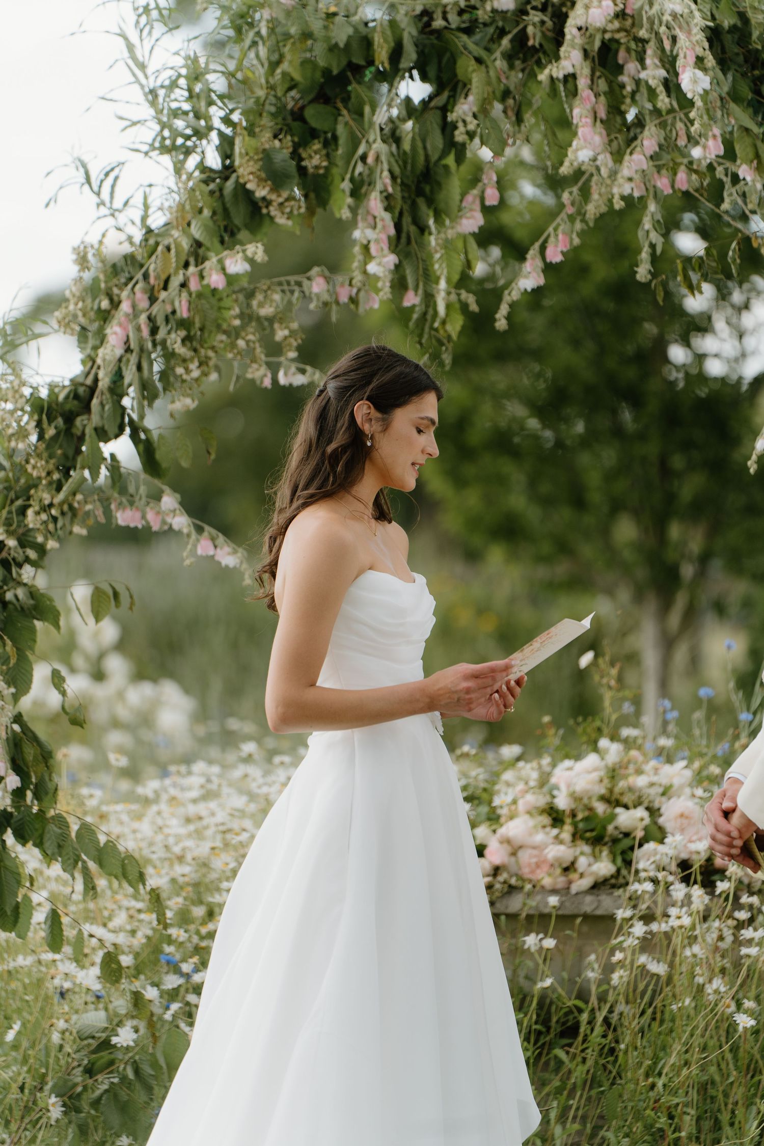 Bride in strapless white gown reading vows under floral arch surrounded by wildflowers.