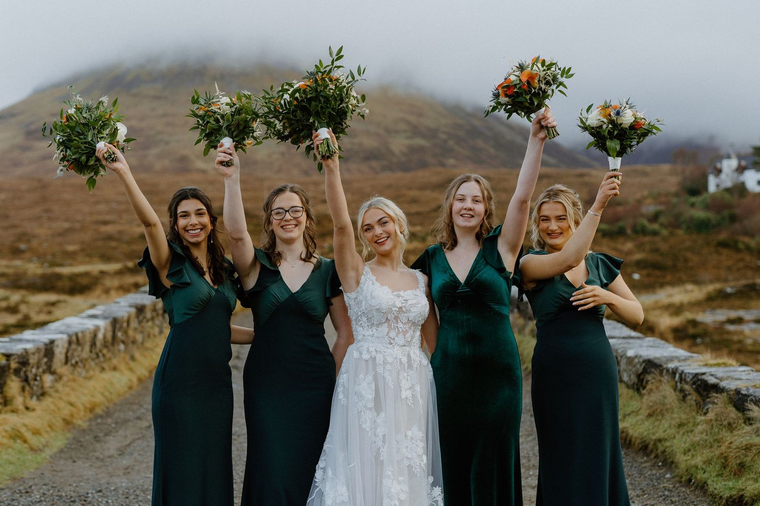 Bride and four bridesmaids in green dresses celebrate raising bouquets before misty Scottish highlands.