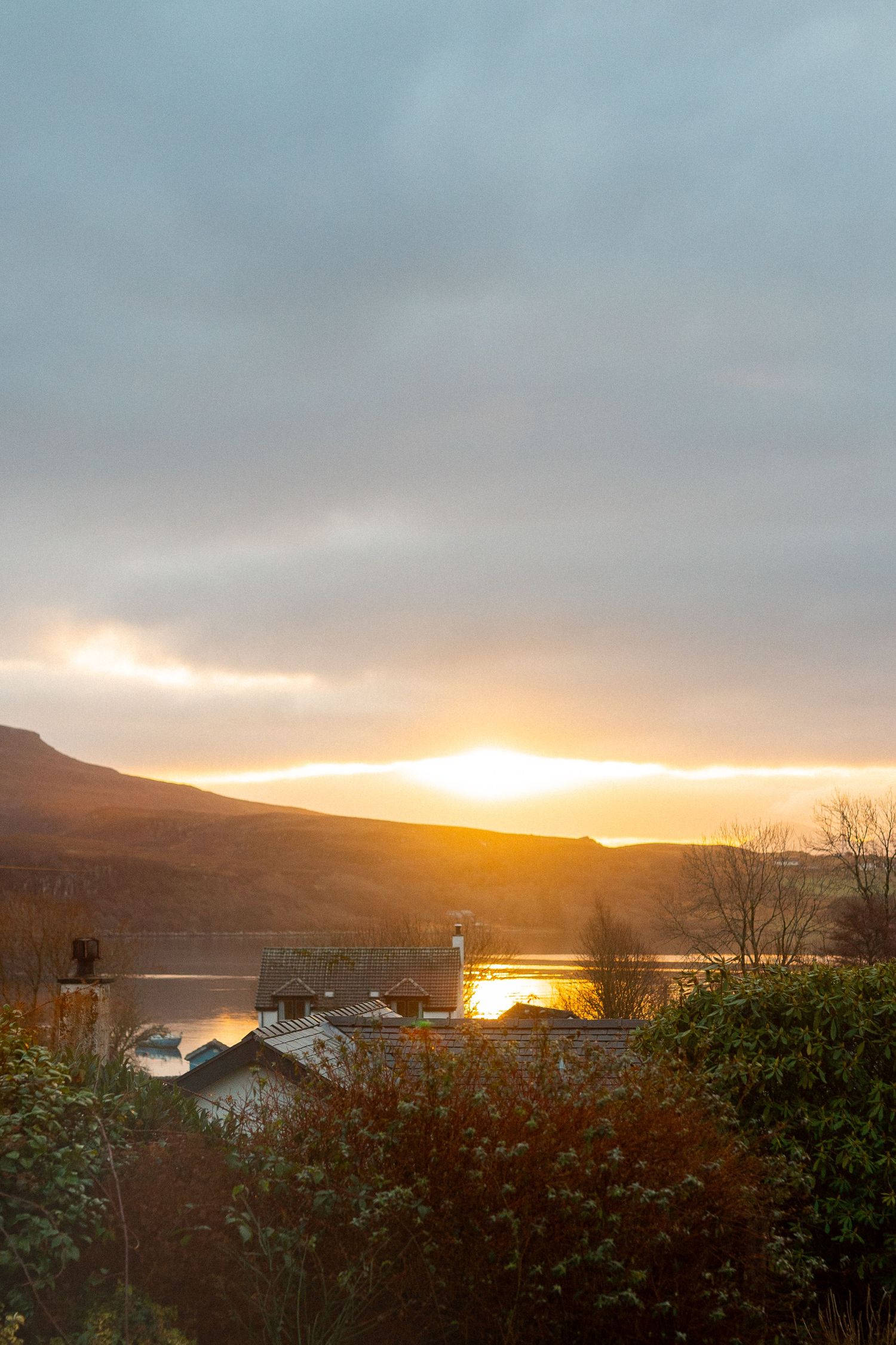 Golden sunset over a loch with rooftops silhouetted against glowing orange sky and rolling hills.