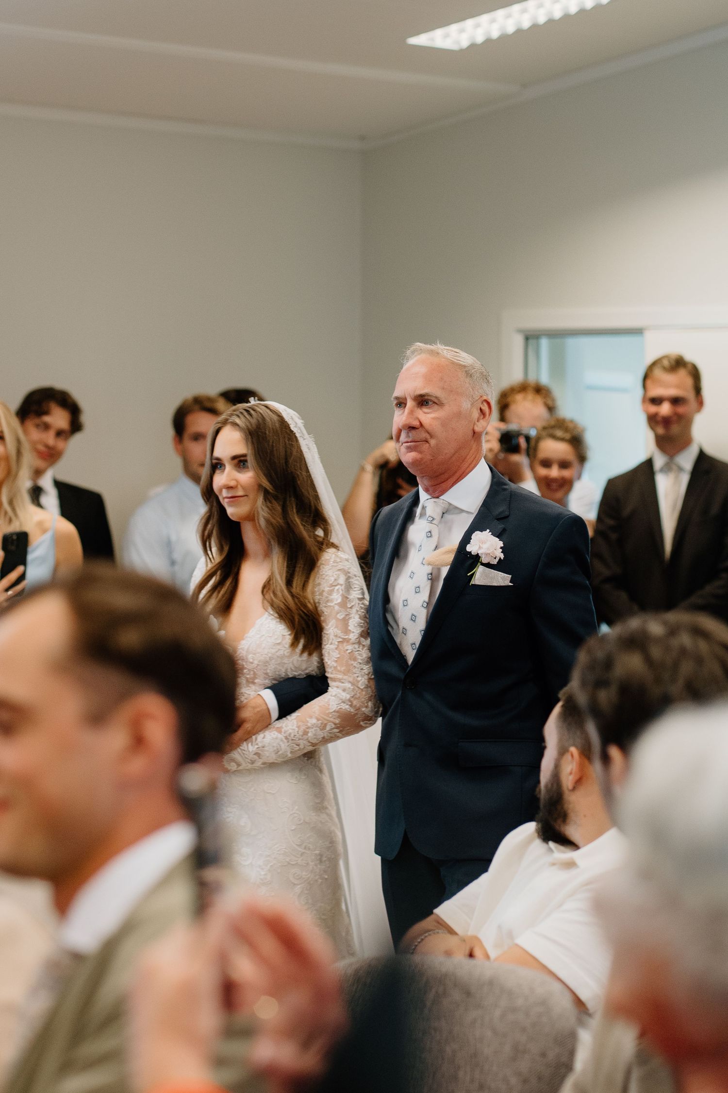 Bride in lace gown with veil walking down aisle escorted by father at wedding ceremony.