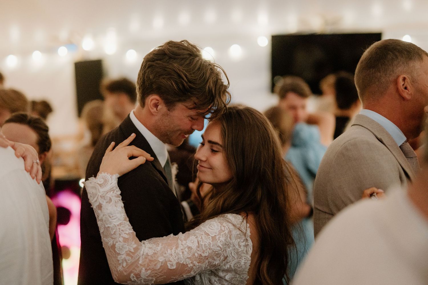 Bride and groom share intimate slow dance embrace under warm string lights at wedding reception.