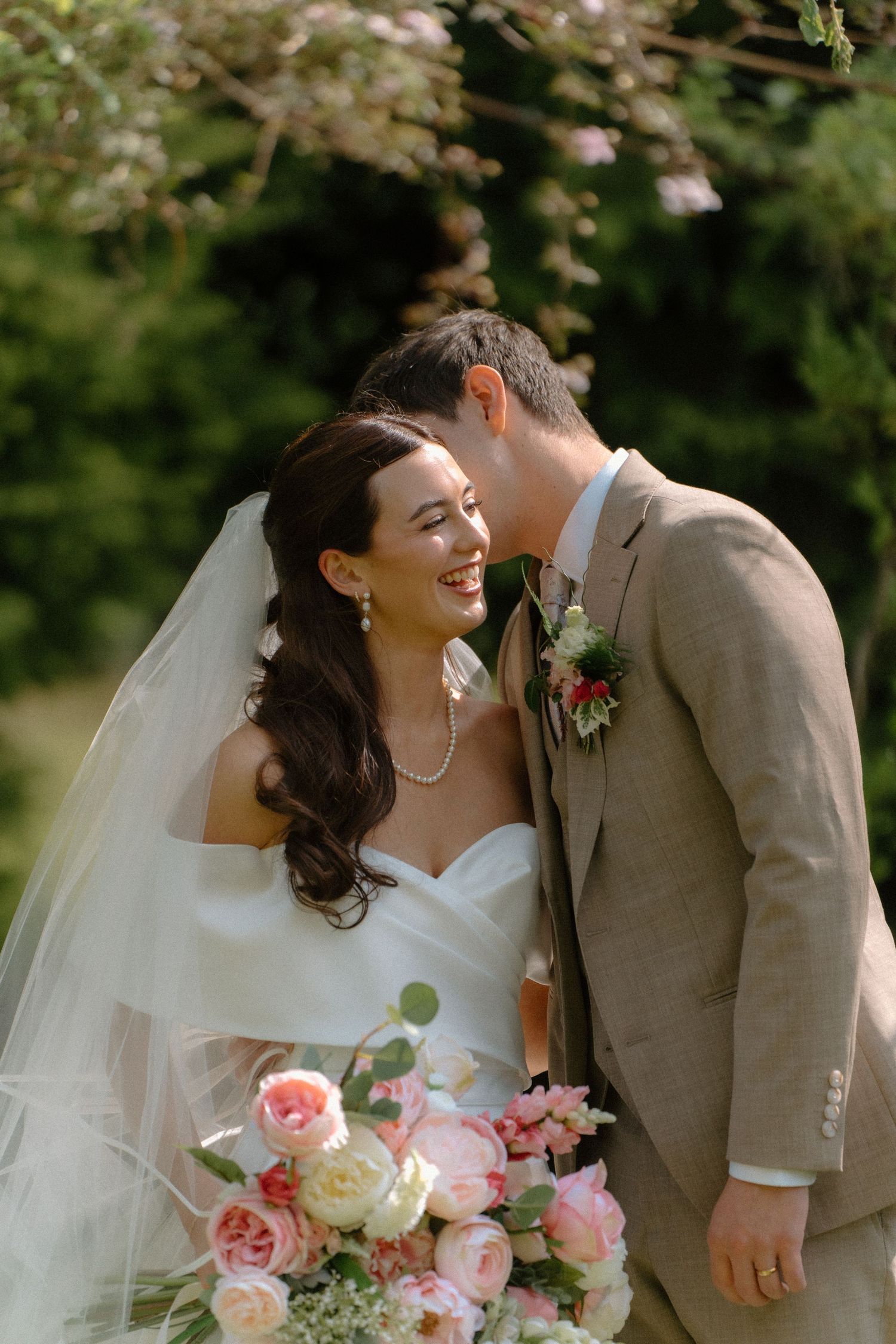 Laughing bride and groom sharing a tender moment surrounded by blooming trees in a garden.