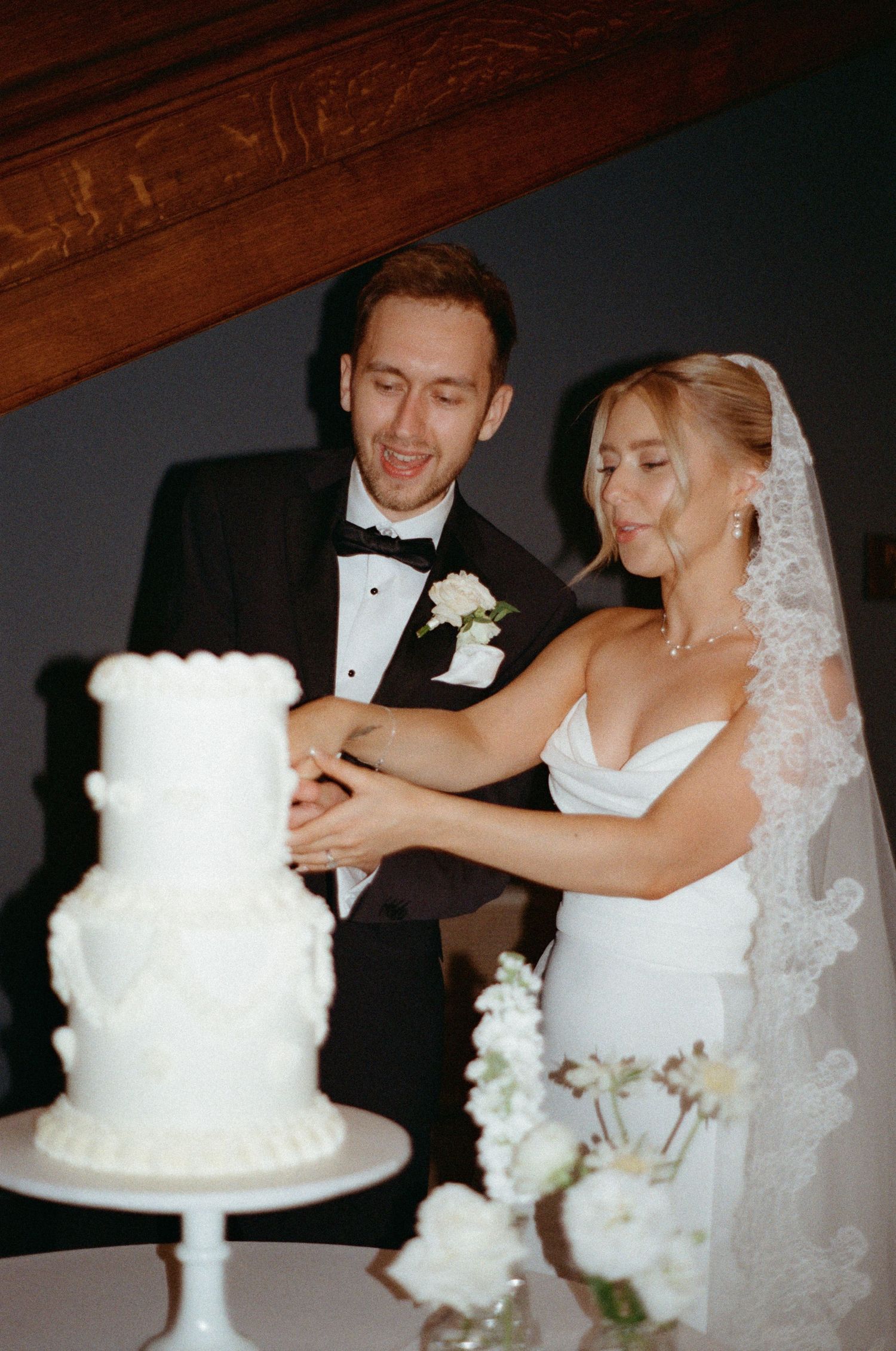 Smiling bride and groom in tuxedo cut elegant three-tier white wedding cake together indoors.