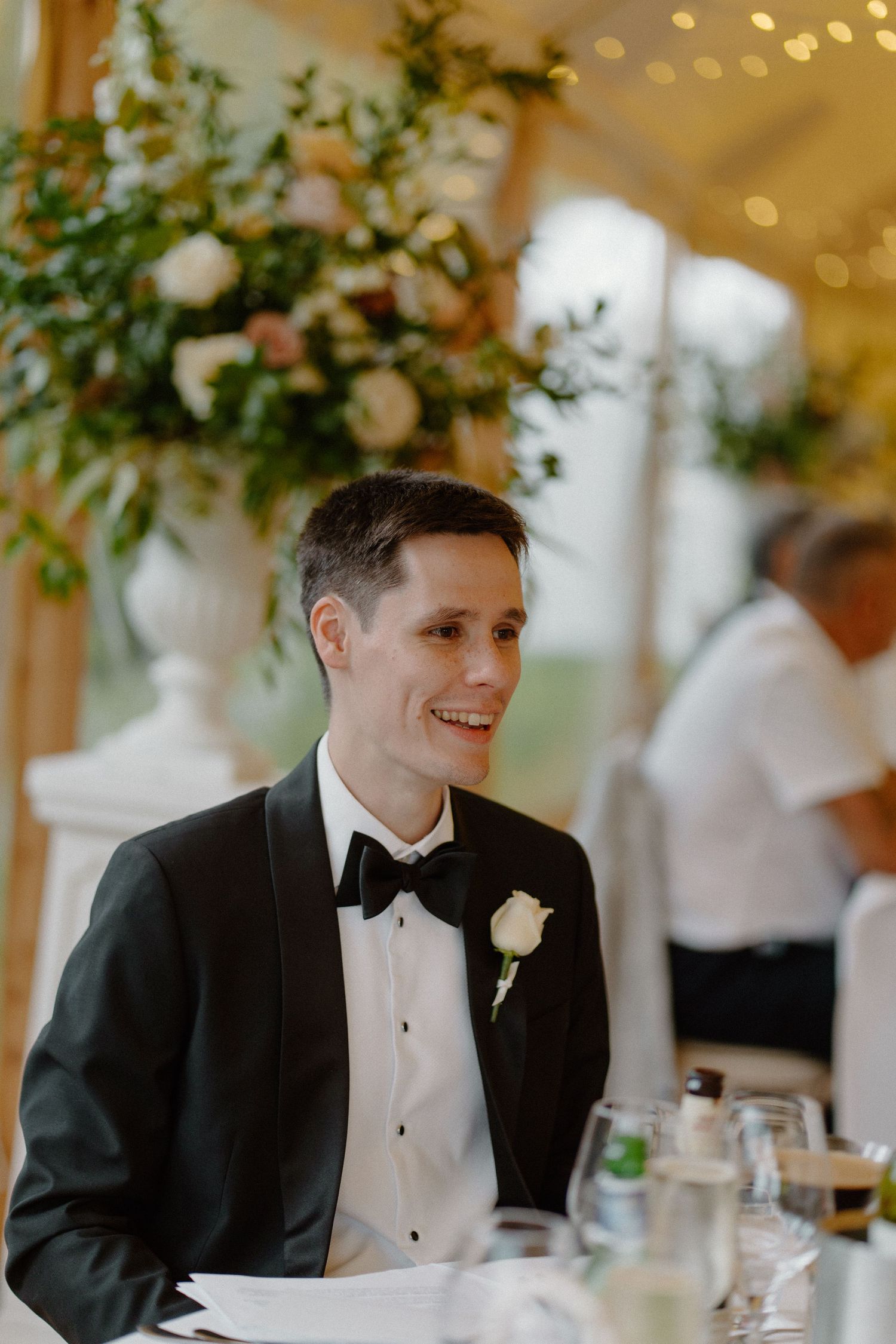 Smiling groom in black tuxedo with white rose boutonniere seated at wedding reception table in marquee.