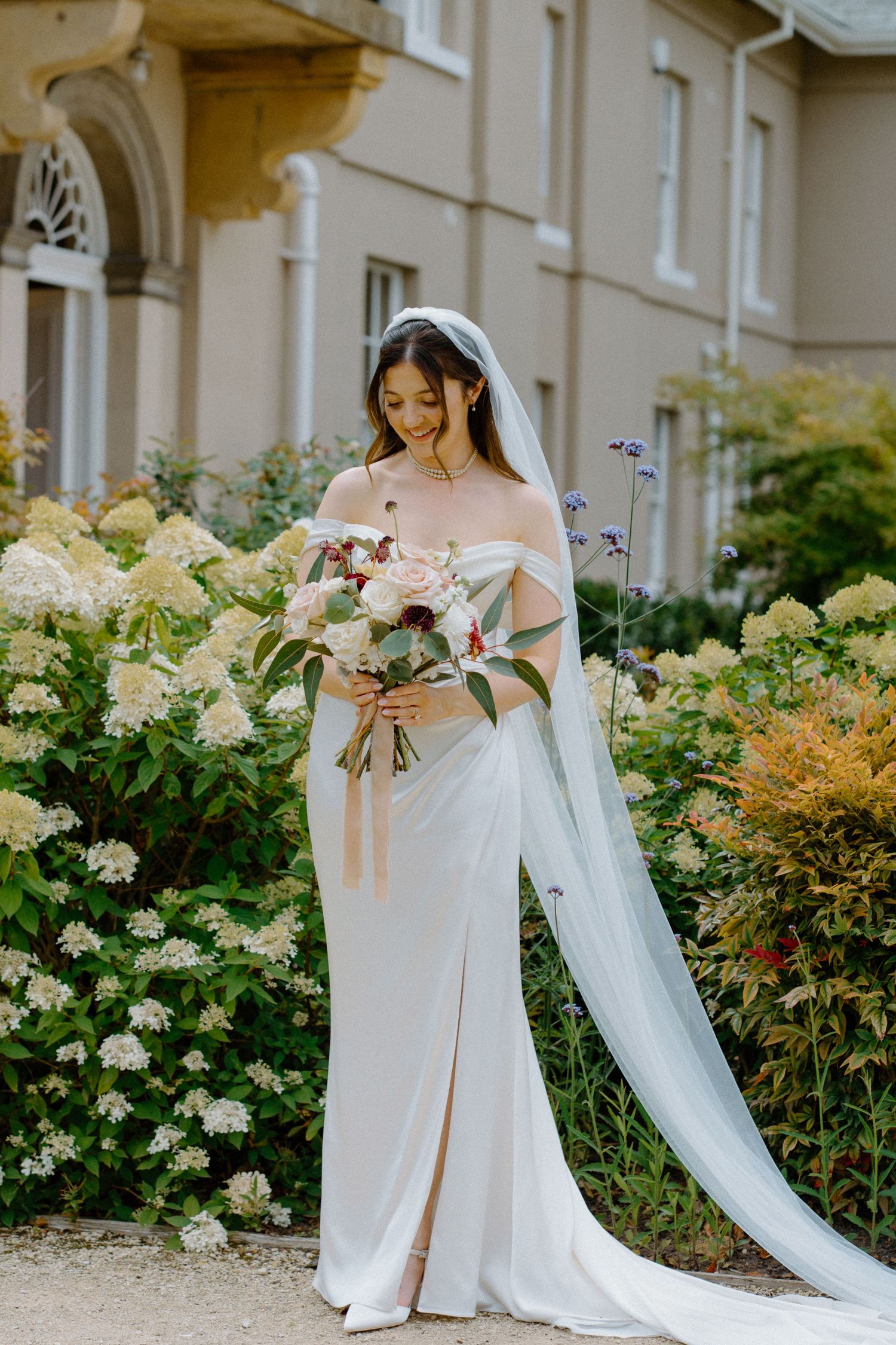 Bride in off-shoulder white gown with veil holding bouquet among hydrangea gardens at elegant venue.