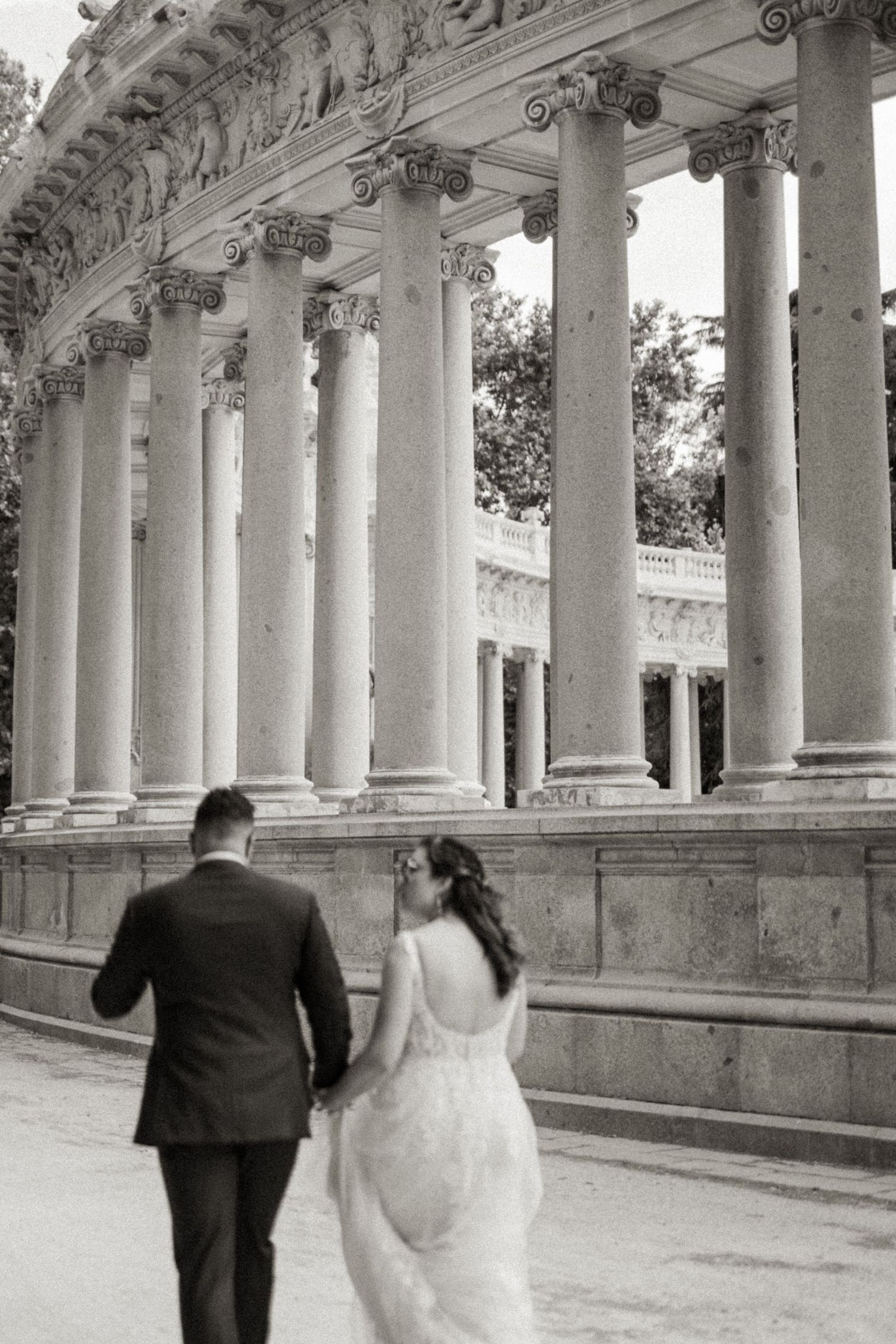 Black and white photo of bride and groom walking past grand classical columns in park.