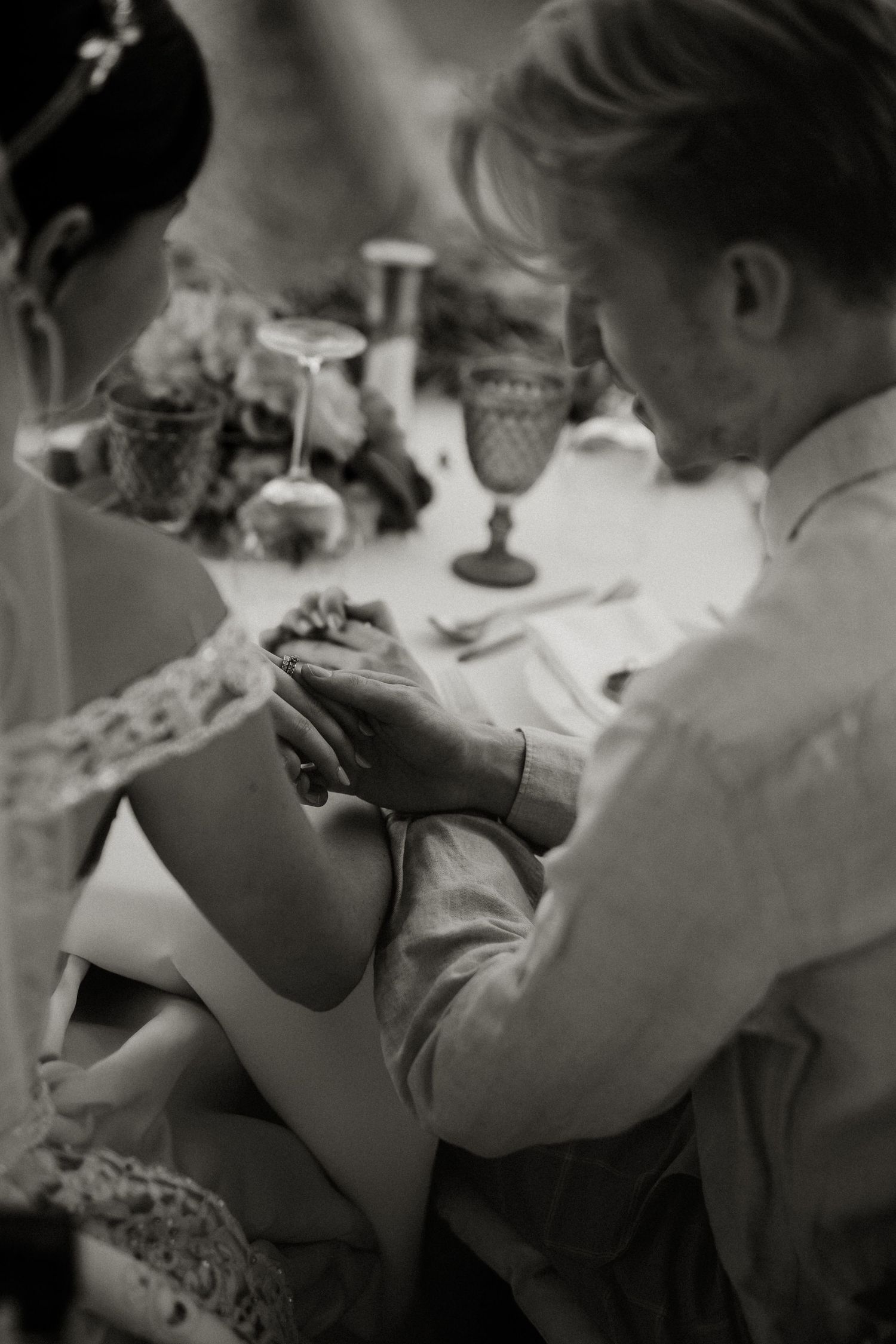 Black and white photo of couple holding hands intimately at wedding reception table.
