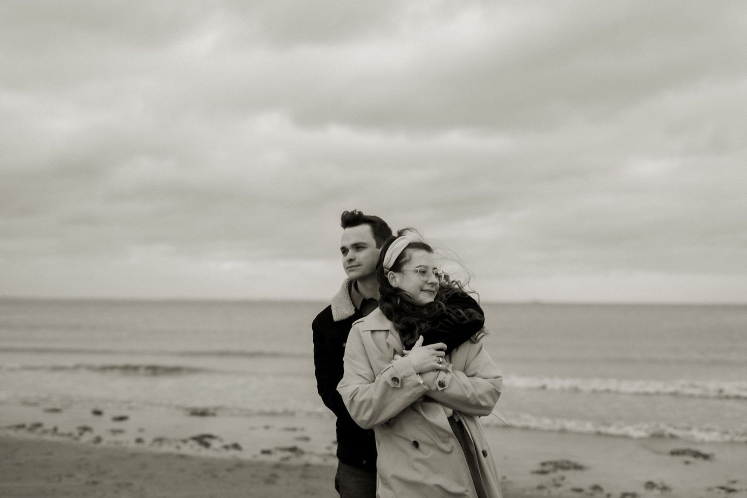 Couple embracing on a beach in black and white, woman leaning into mans arms.