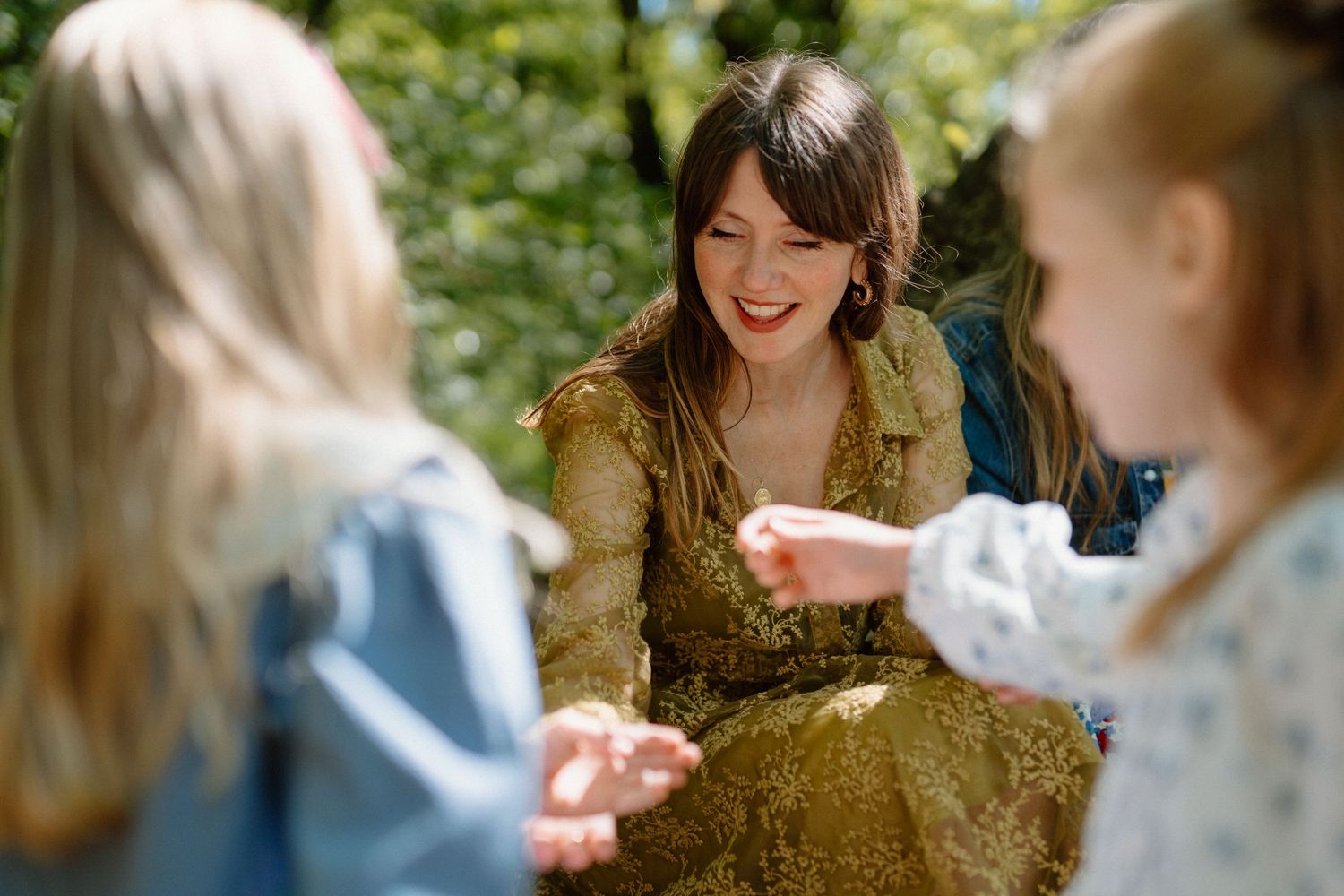 Smiling brunette woman in gold dress interacting joyfully with two young girls outdoors.