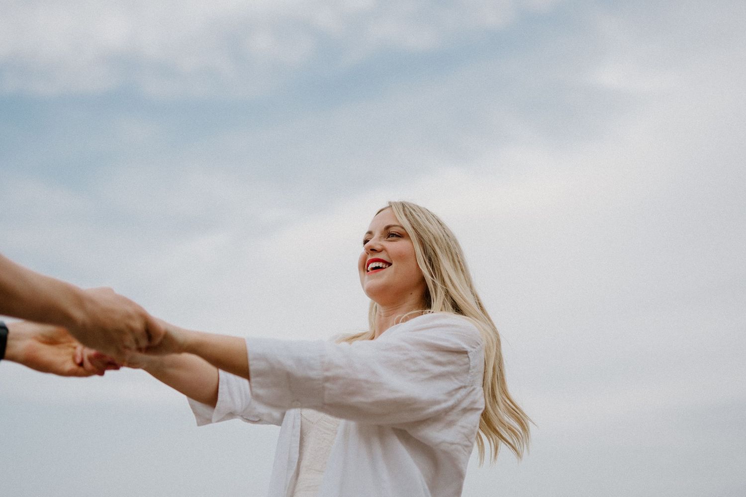 Smiling blonde woman in white shirt holding hands with partner against a cloudy sky backdrop.