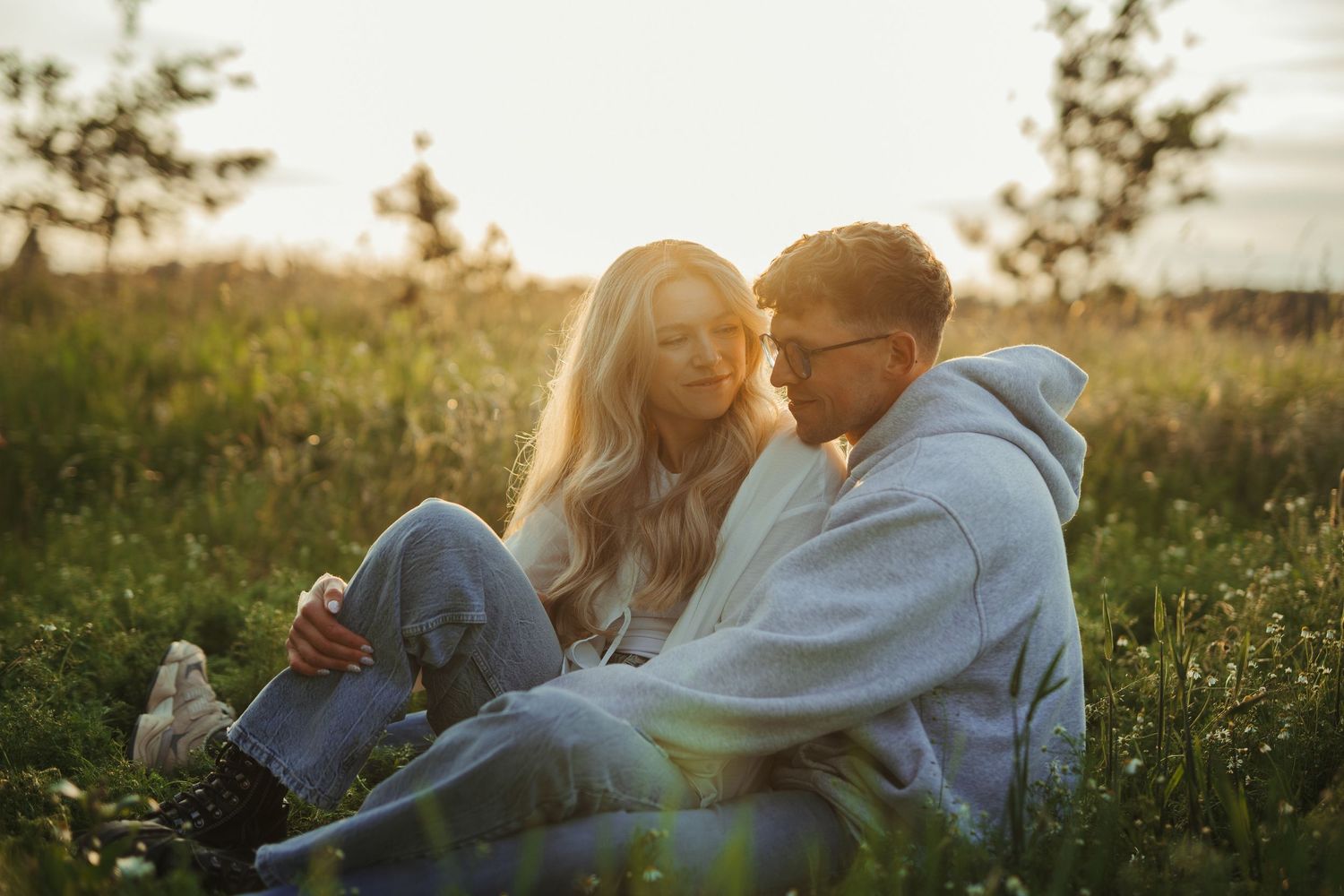 Couple sitting together in green meadow at sunset, woman leaning on mans shoulder in warm light.