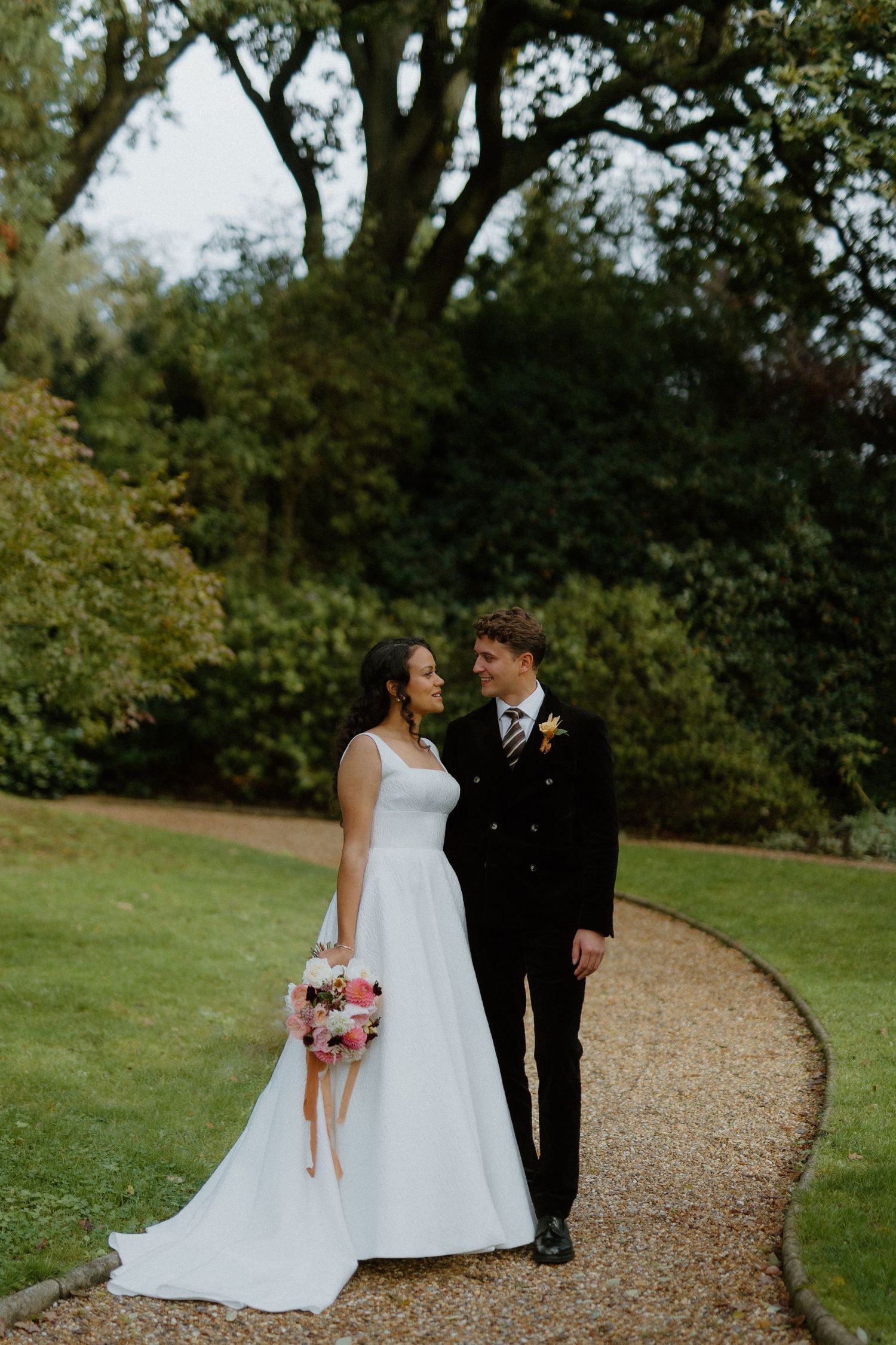 Bride and groom walking hand in hand along curved garden path surrounded by lush green trees.