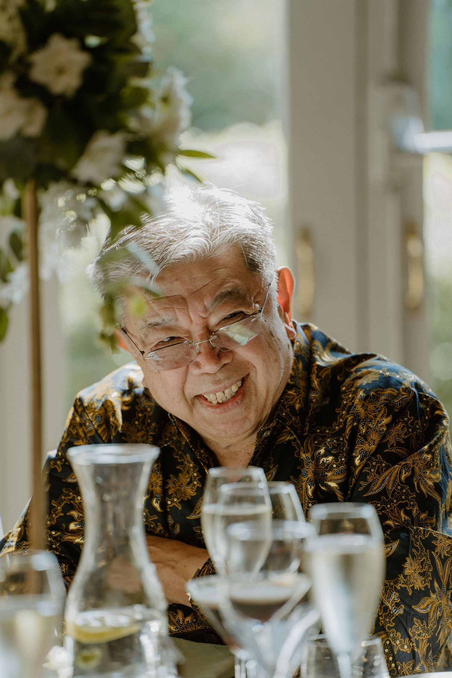 Smiling elderly man in floral shirt enjoying wedding reception with champagne glasses in foreground.