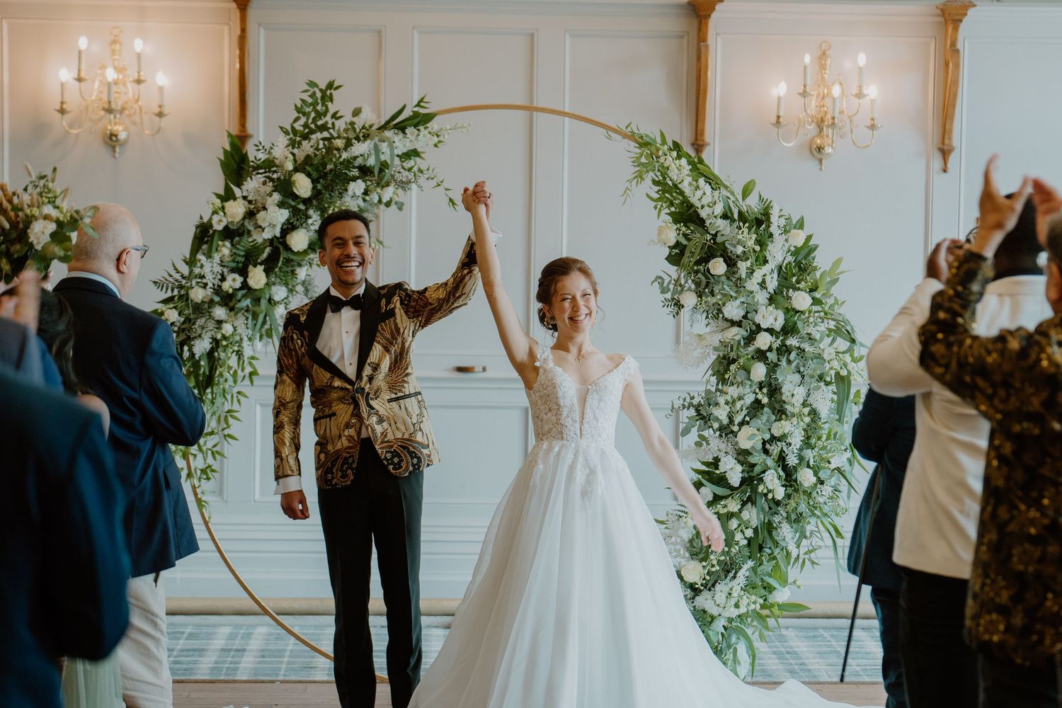 Newlyweds celebrate recessional under gold hoop floral arch in elegant wedding venue.