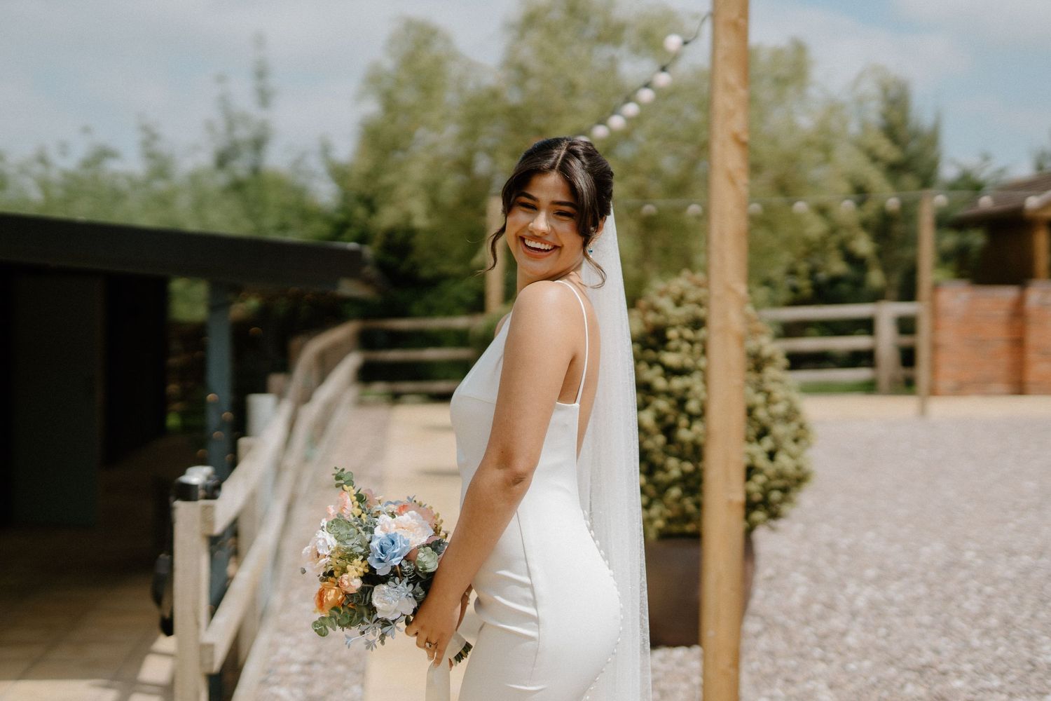 Bride in fitted white dress and veil glancing back with a smile while holding a blue and peach bouquet.