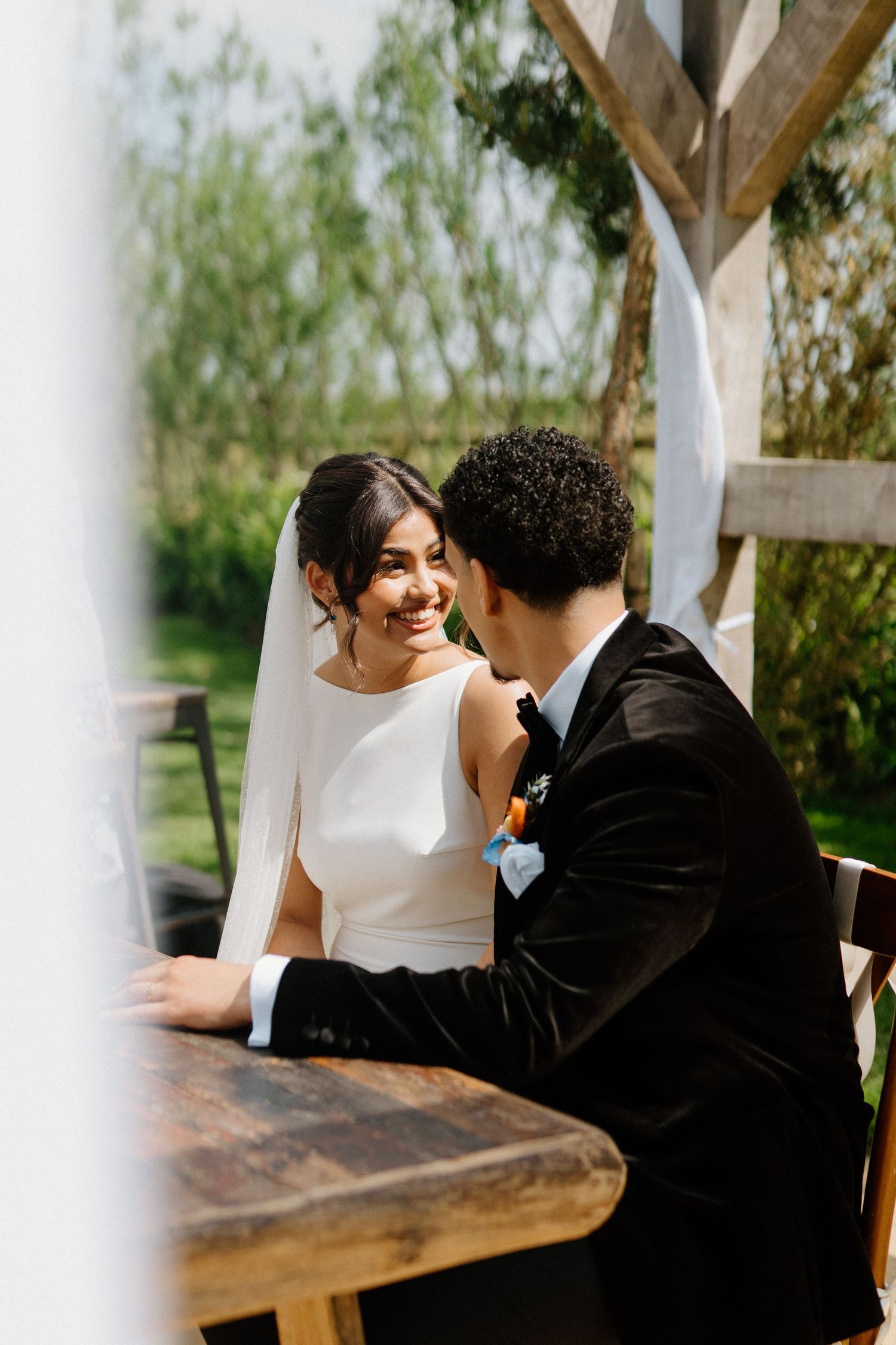 Radiant bride in white gown with veil smiles at groom in black tuxedo while seated at rustic wooden table.