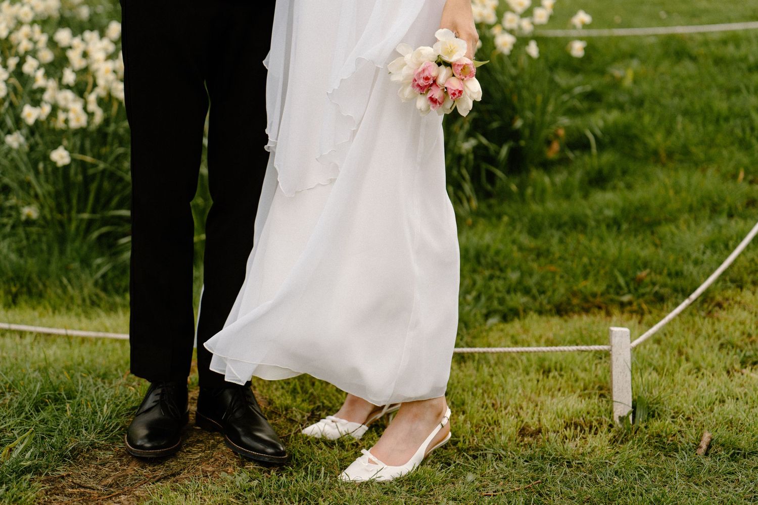 Close-up of bride and groom's feet, bride wearing white flats holding pink bouquet, groom in black dress shoes on green grass.