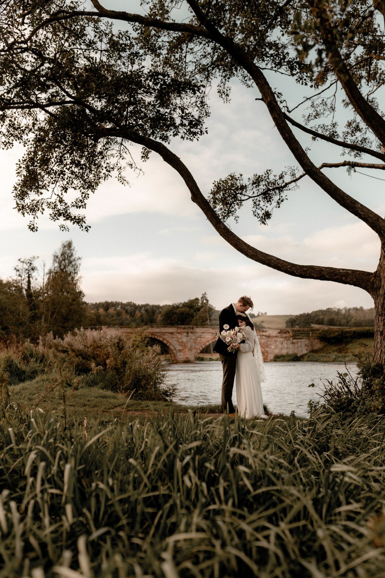 A dreamy wedding portrait under arching tree branches beside a tranquil river.