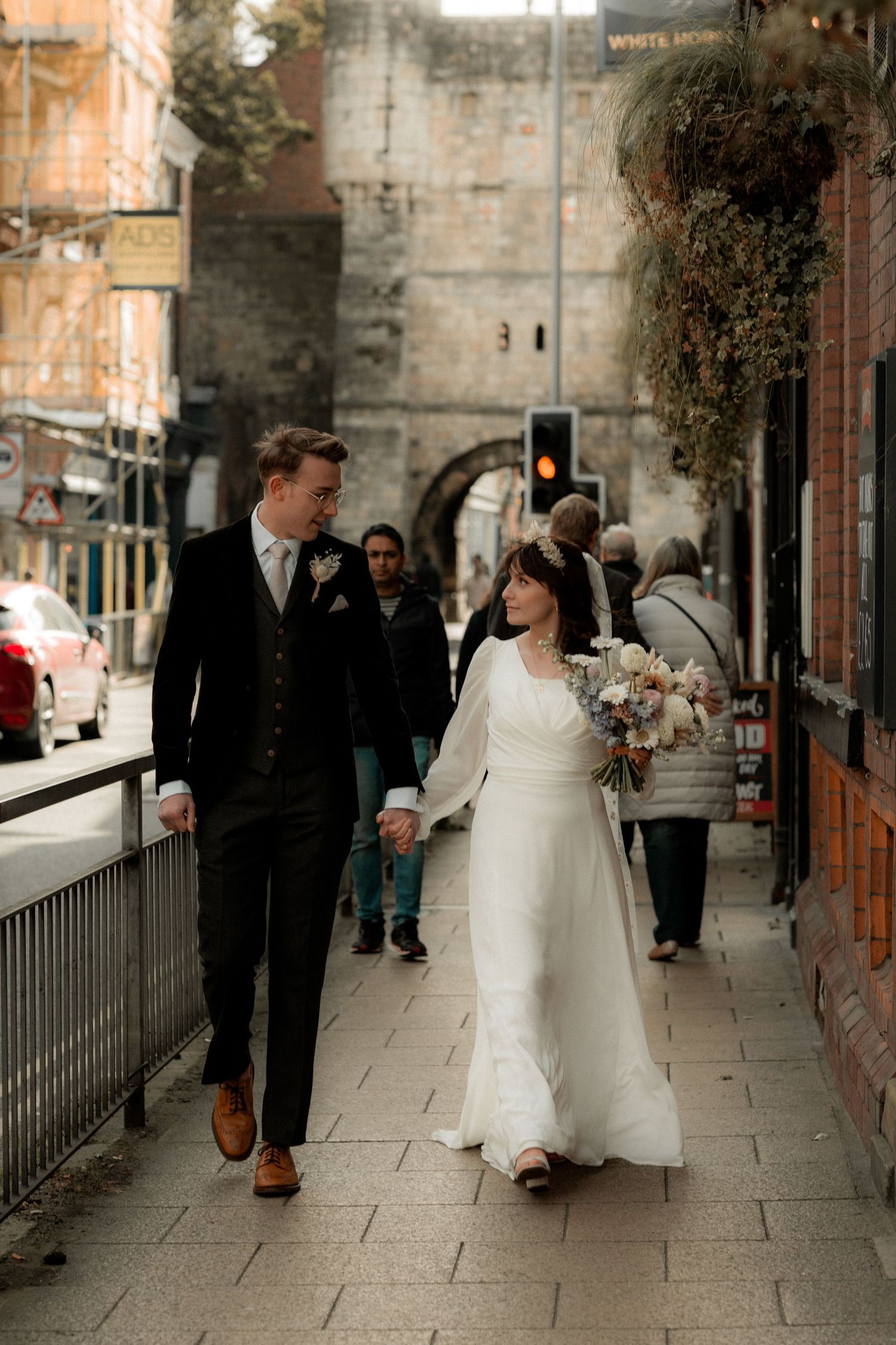 A couple in wedding attire walking along a historic stone street with ancient walls in the background.