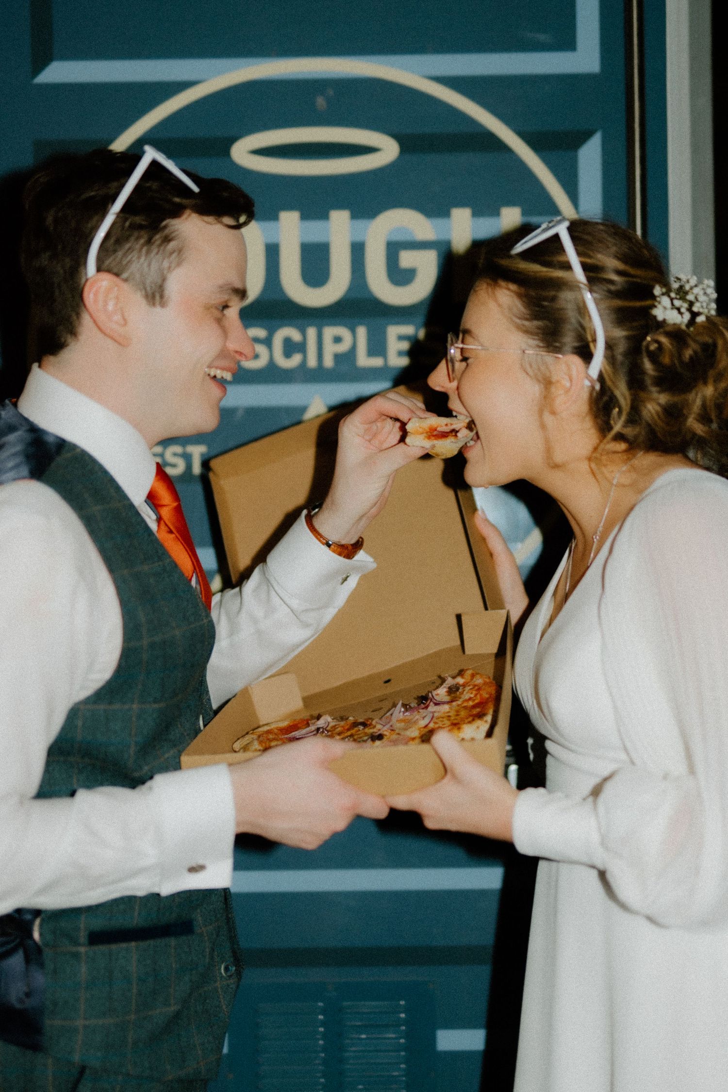 A joyful couple shares pizza at what appears to be their wedding reception, with a blue neon sign visible in the background.