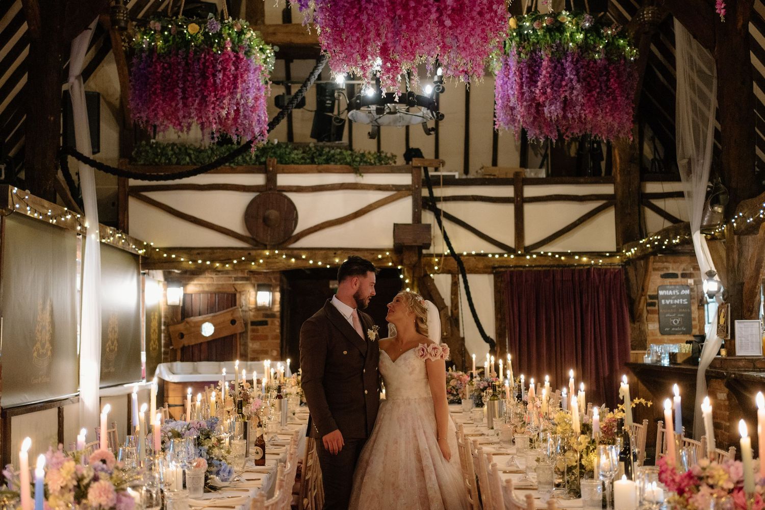 Elegant wedding reception venue decorated with hanging pink wisteria, string lights and candlelit tables for evening celebration.