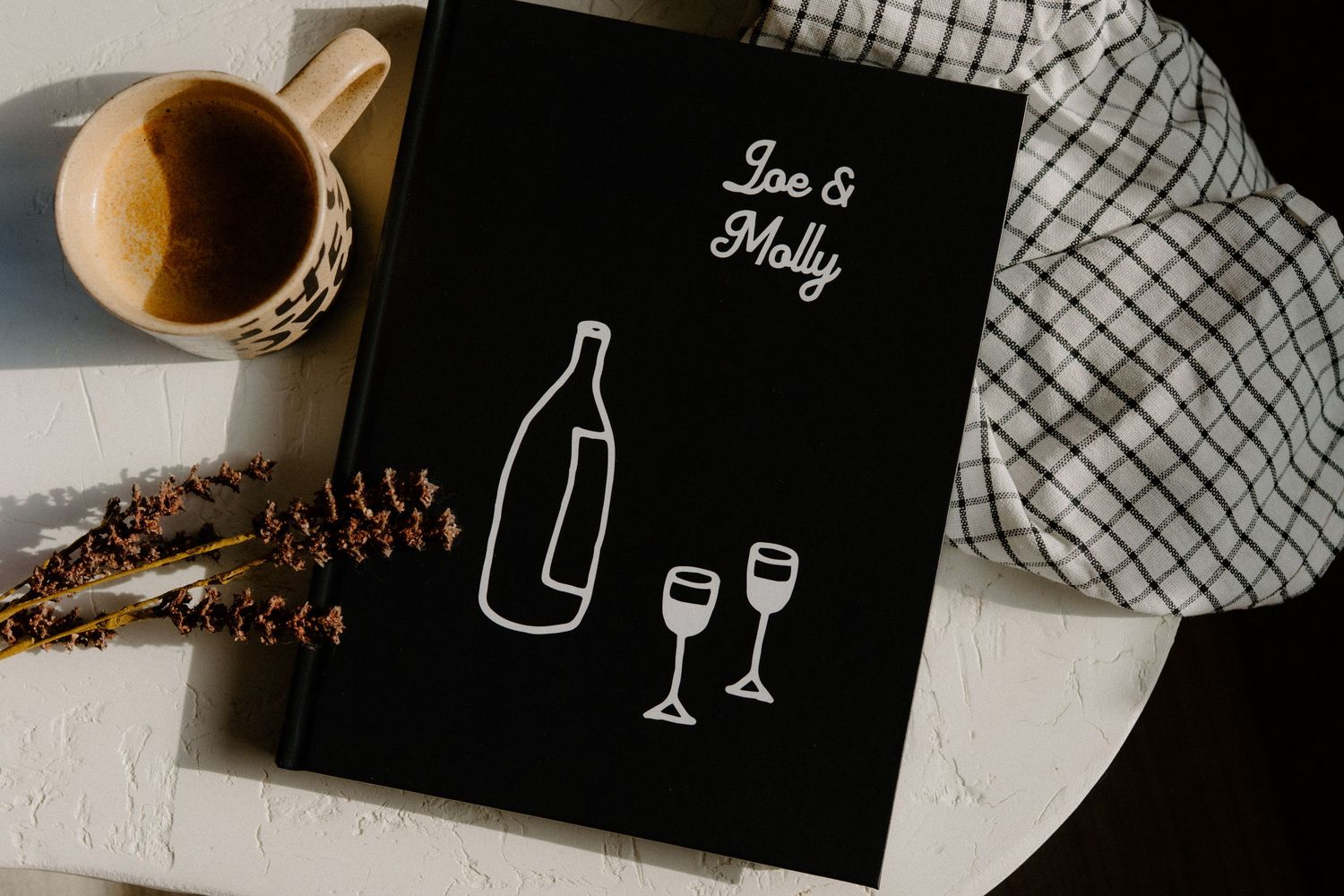 A black book with wine bottle illustration lies next to a coffee cup and pinecone on a white surface with checkered cloth.