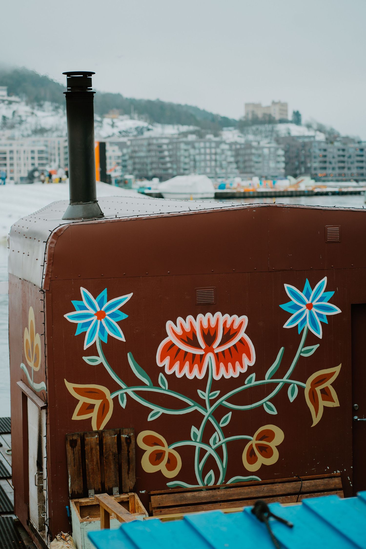 A brown container with decorative floral art featuring blue and orange flowers sits near a waterfront cityscape.