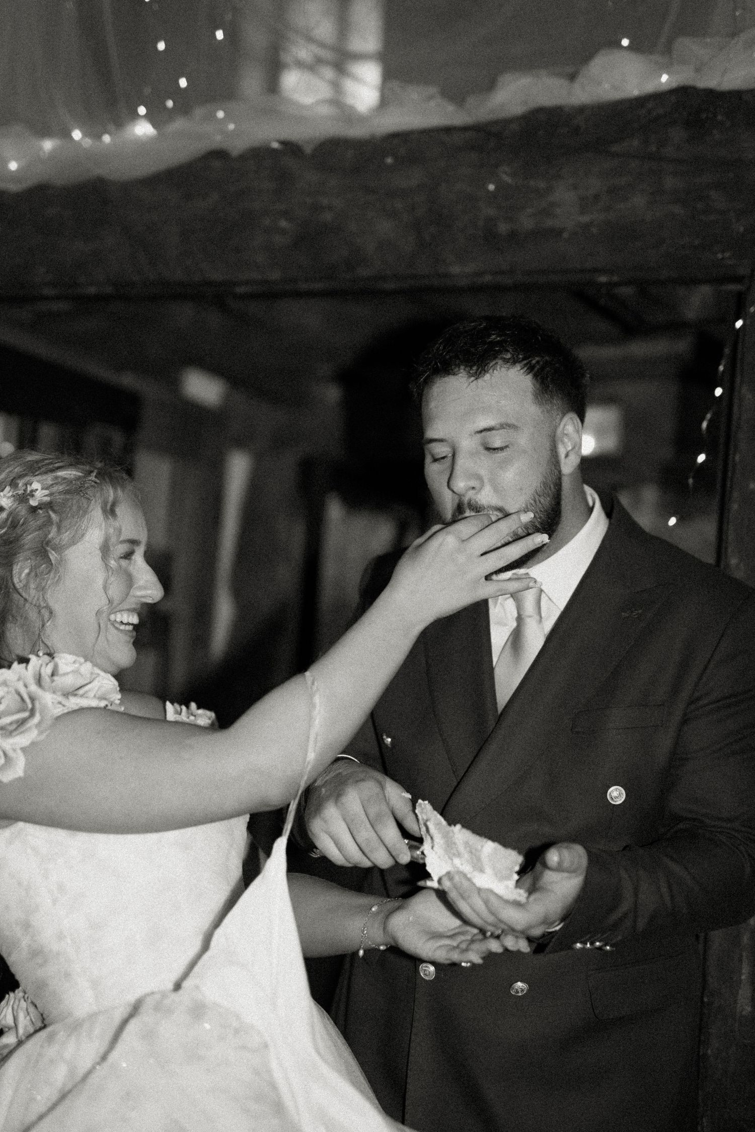 Black and white photo of laughing bride feeding wedding cake to groom in a rustic barn setting.