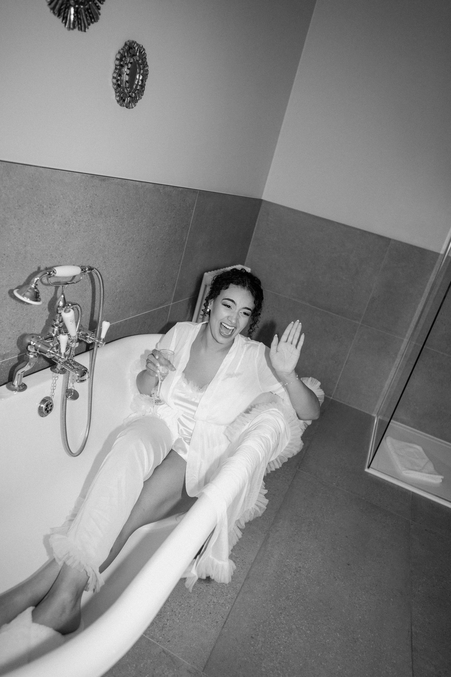 Bride in white robe sitting in a clawfoot bathtub holding champagne, black and white photo.