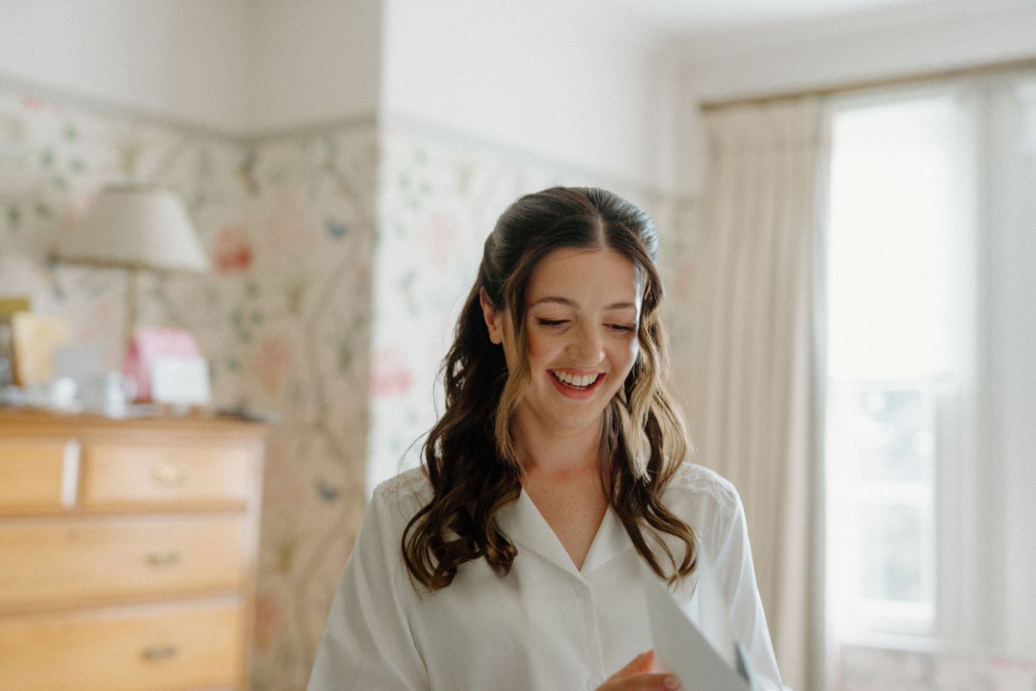 Bride in white robe smiling while reading a letter in a bright bedroom.