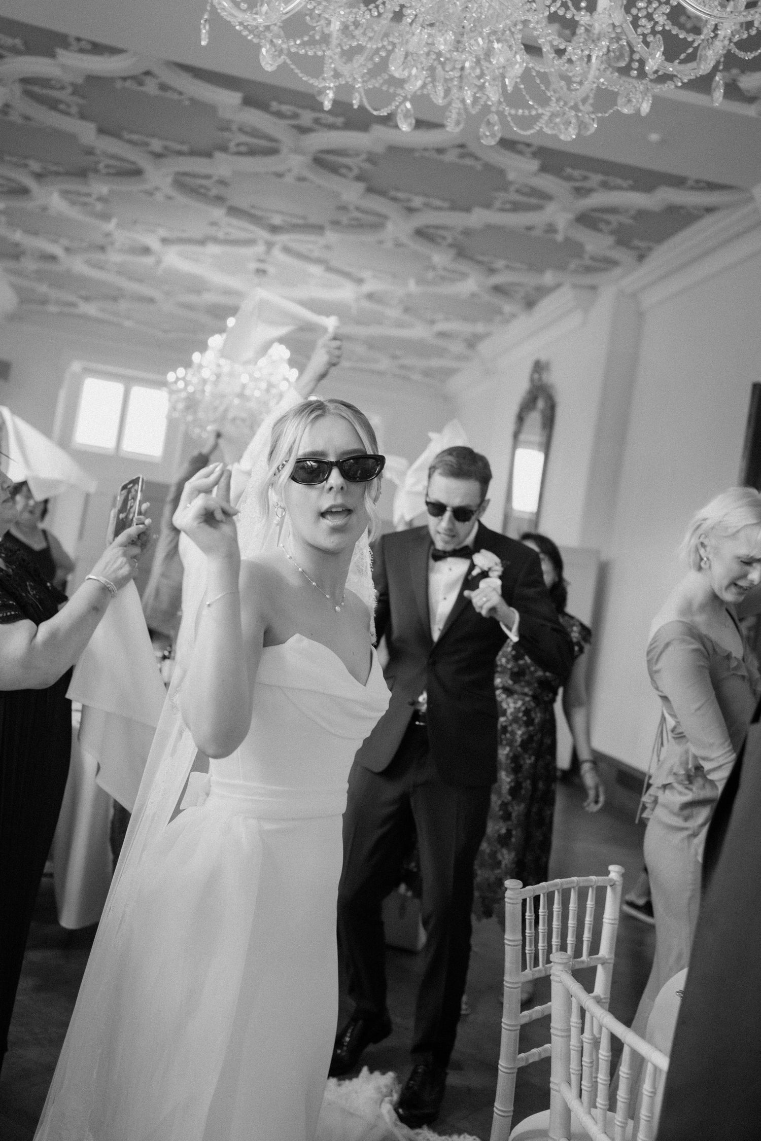 Black and white photo of bride in sunglasses and groom making a fun entrance into their wedding reception.